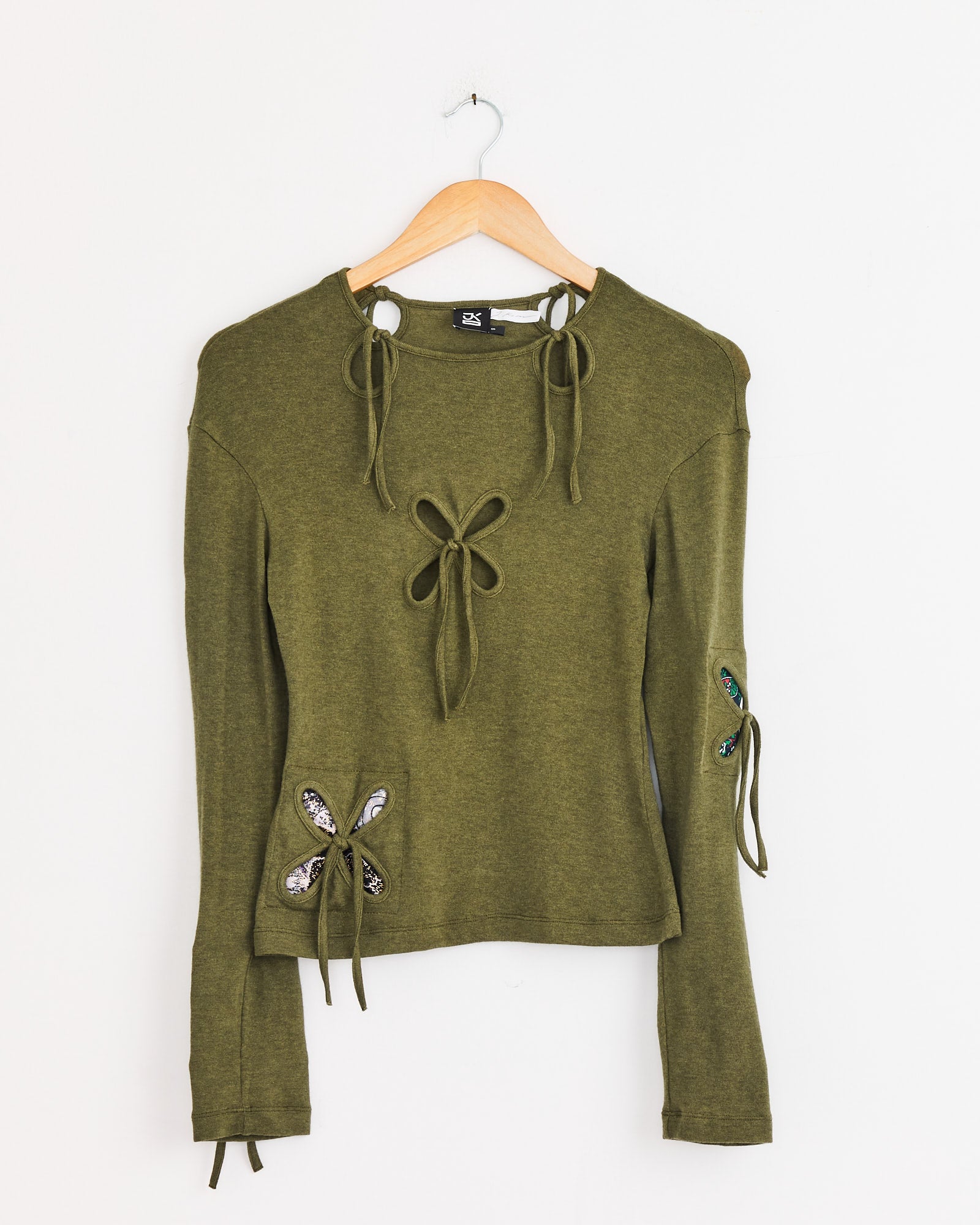 The J. Kim Petal Patches Top in Swamp Green features long sleeves and tie details at the neckline, front, sleeves, and lower side. Displayed on a wooden hanger against a plain white background.