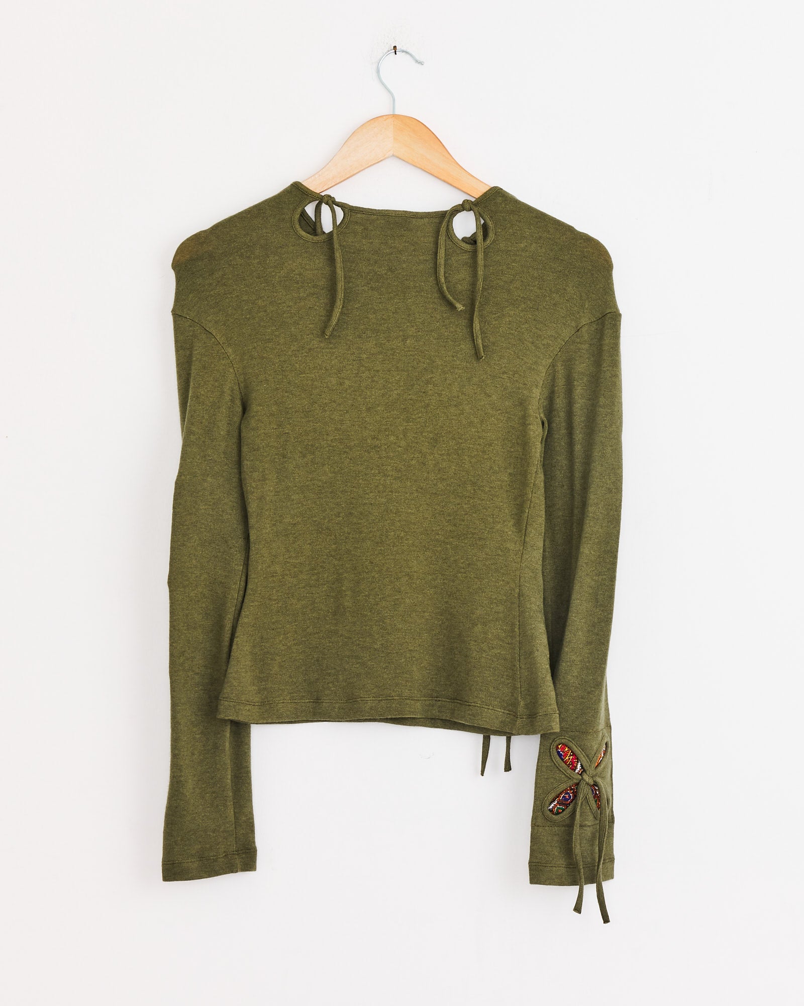 The Petal Patches Top in Swamp Green by J. Kim hangs on a wooden hanger, featuring long sleeves, ties at the neckline and sleeves, and a small red and black embroidered patch on one sleeve.