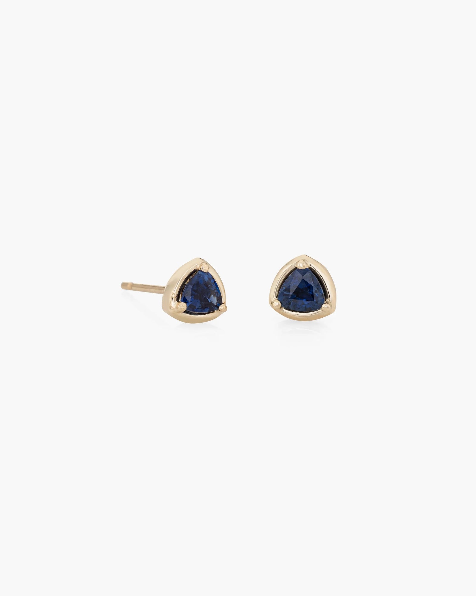 The Prism Studs Pair by Kathryn Bentley features deep blue, triangular sapphires set in 14K yellow gold prongs, displayed against a white background.