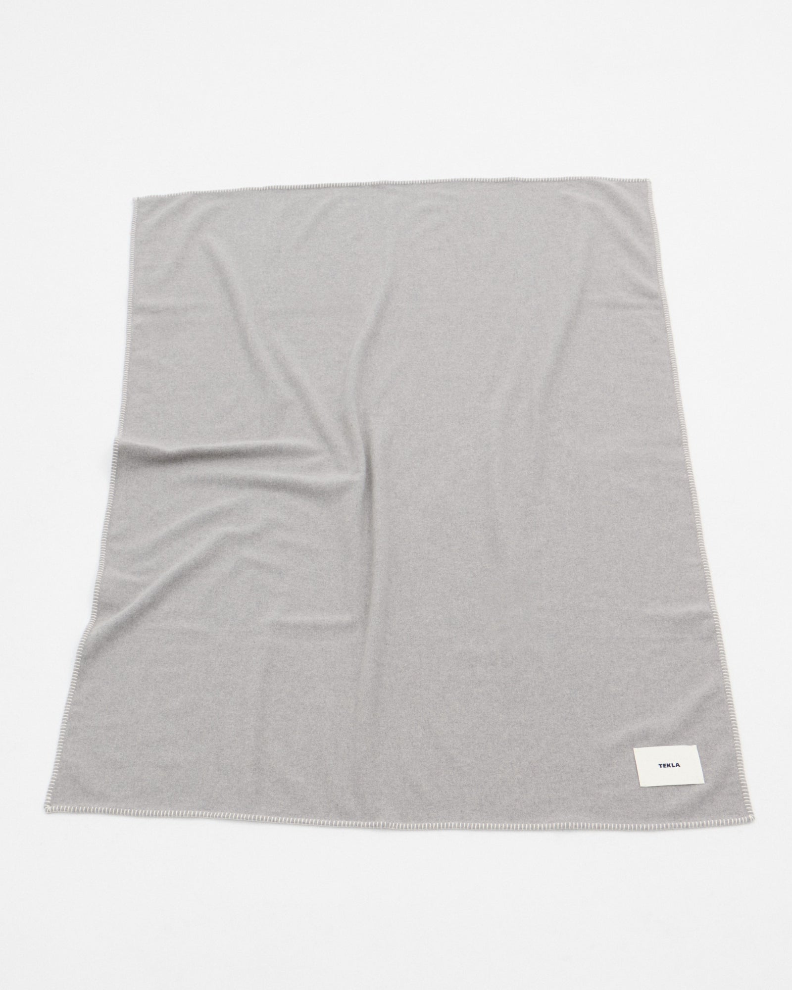 The TEKLA Lambswool Blanket in Ash, a light gray rectangular piece with a small white label in one corner, is made in Lithuania and displayed flat on a white surface with subtle wrinkles.