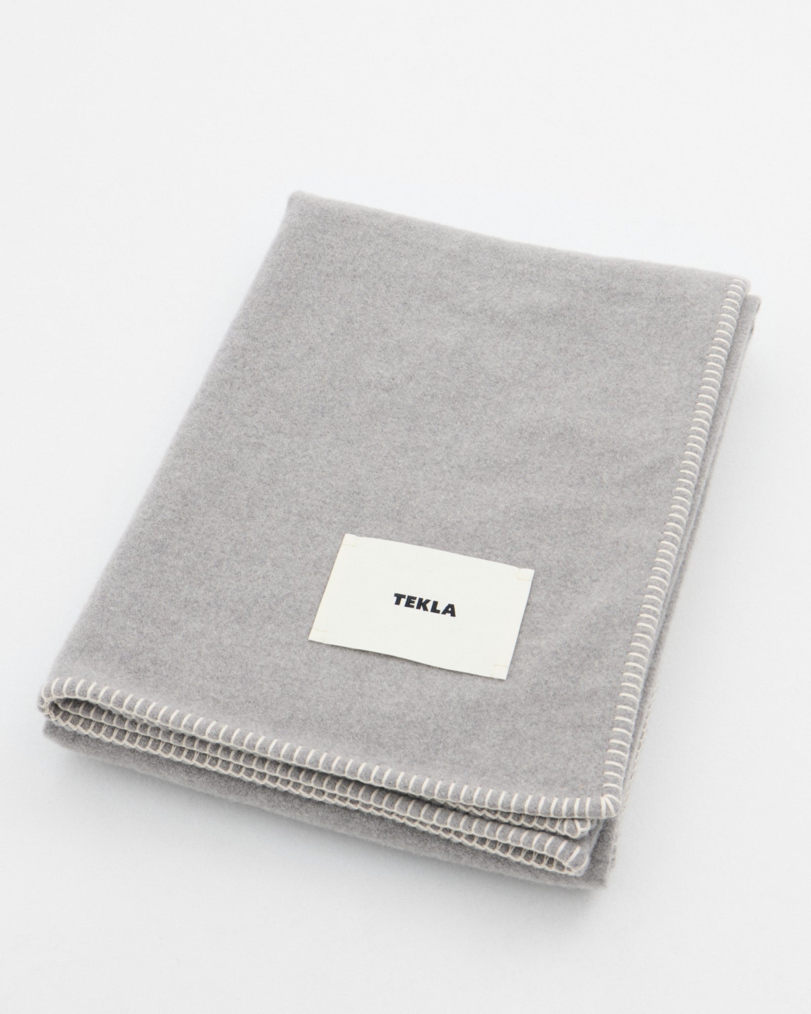 The TEKLA Lambswool Blanket in Ash, made in Lithuania, features a light gray color, white stitched edges, and a rectangular TEKLA label. It is shown folded on a plain white surface.