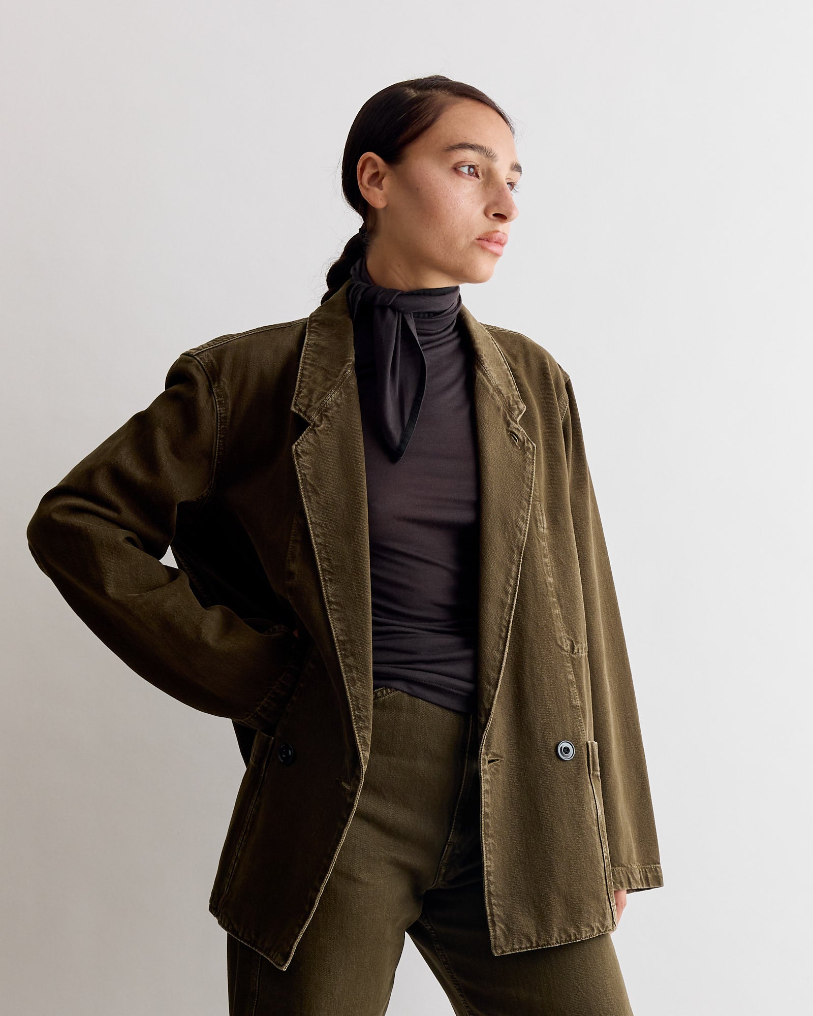 A woman stands against a plain background in a Lemaire Foulard Patches Top in Coal, paired with an olive green blazer and pants. She looks to the side with one hand on her hip, her hair pulled back.