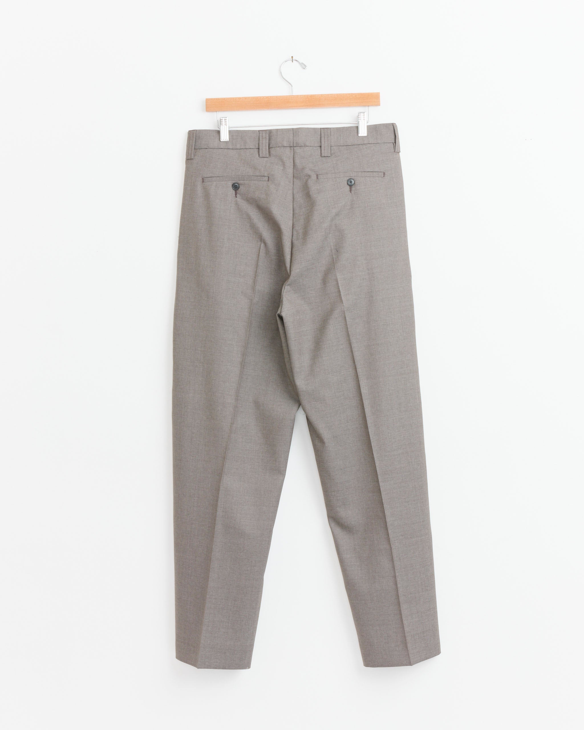 The mfpen Classic Wool Trouser in Light Taupe, crafted from 100% wool in PT, hangs on a wooden hanger against a white background. It features two back pockets with button closures and belt loops at the waistband.