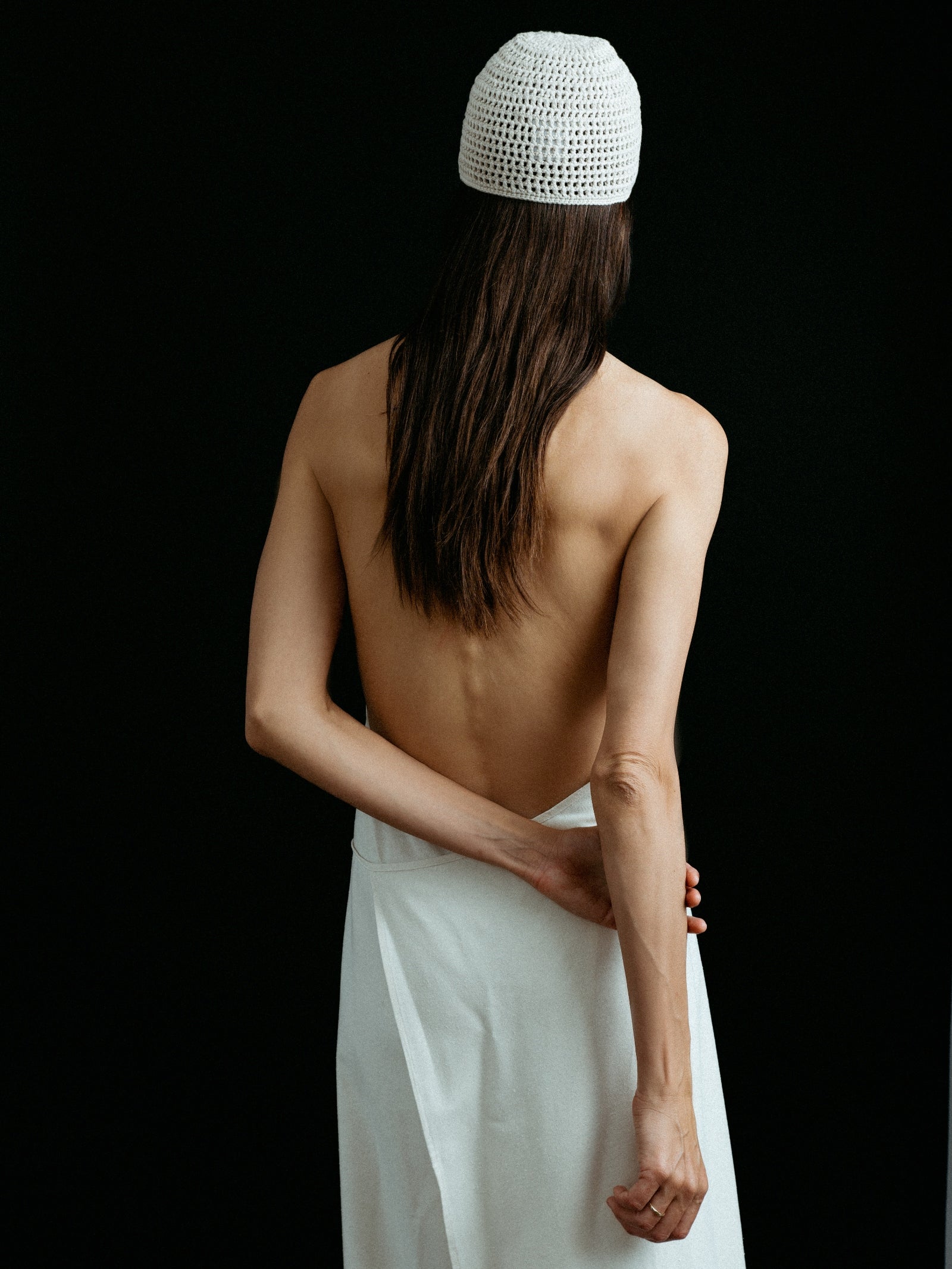 A person with long brown hair, seen from behind, wears the Baserange Apron Dress in Off White, revealing their bare back and arm against a black background.