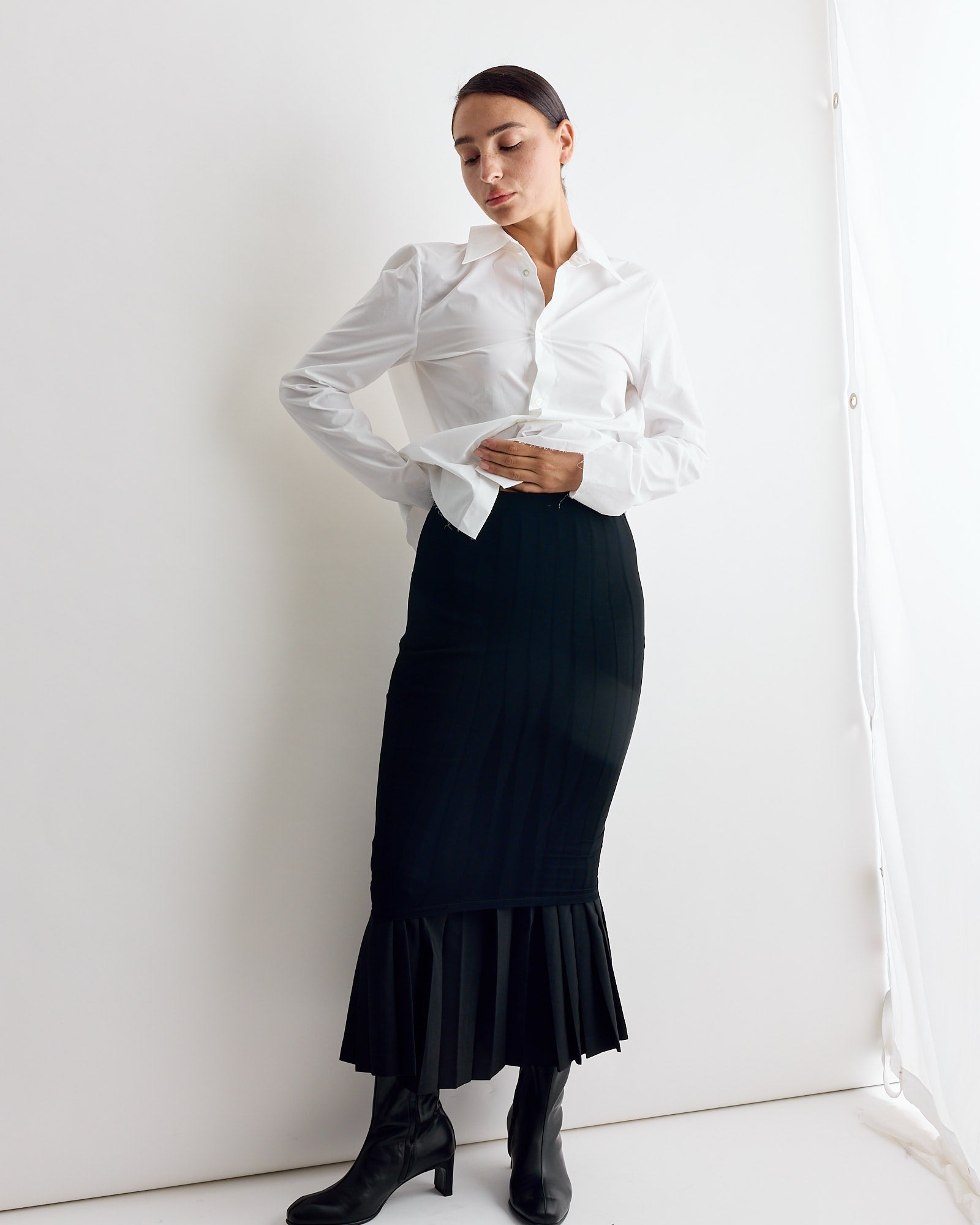 A person stands against a white wall, wearing a white collared shirt, black boots, and the MM6 Maison Margiela Long Skirt IN Black—a structured pleated skirt with a fitted silhouette—looking down with hands near their waist.