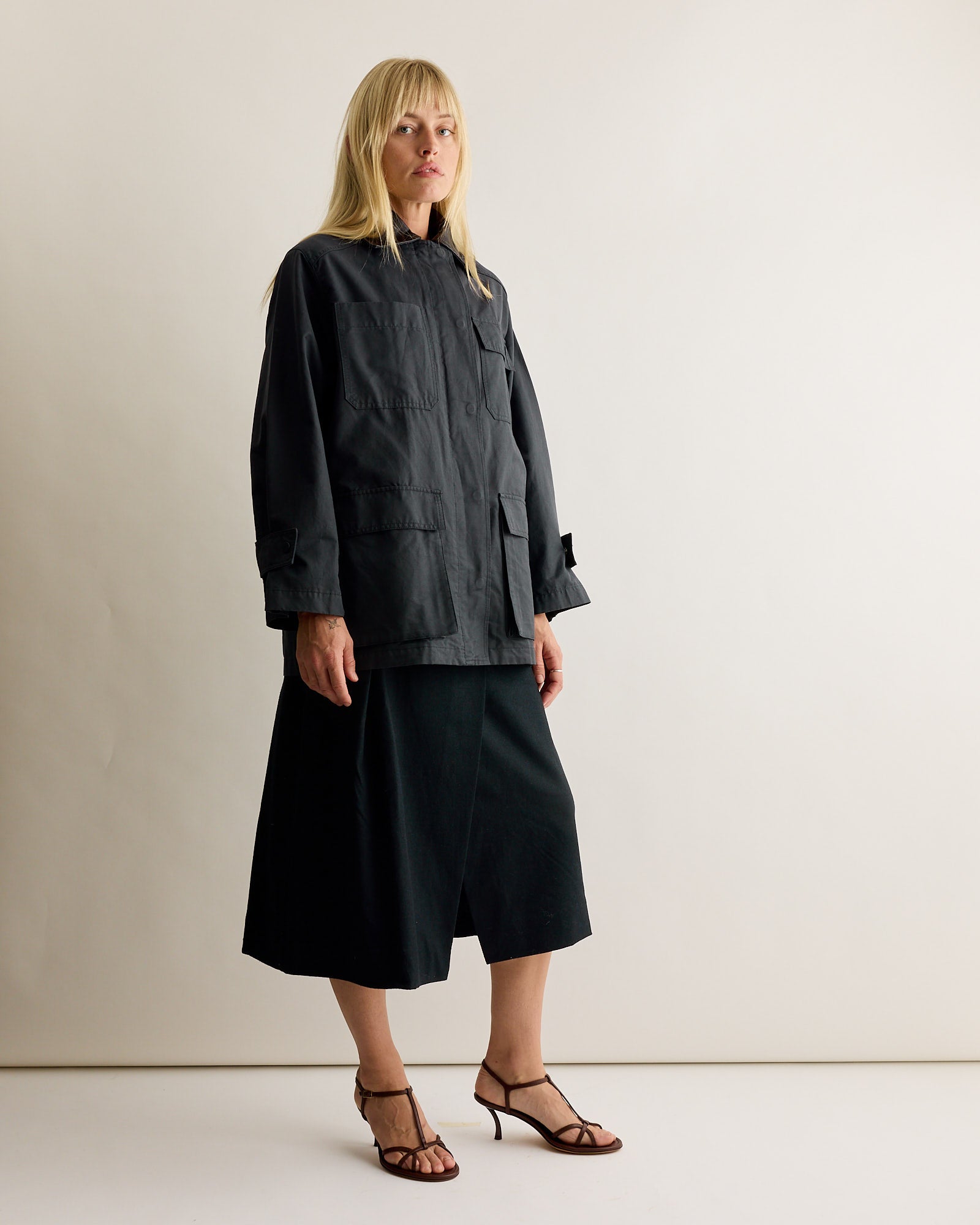 A woman with straight blonde hair and a neutral expression stands against a plain light background, wearing the Mijeong Park Contrast Collar Work Jacket in Navy, a dark midi skirt, and black strappy heeled sandals.