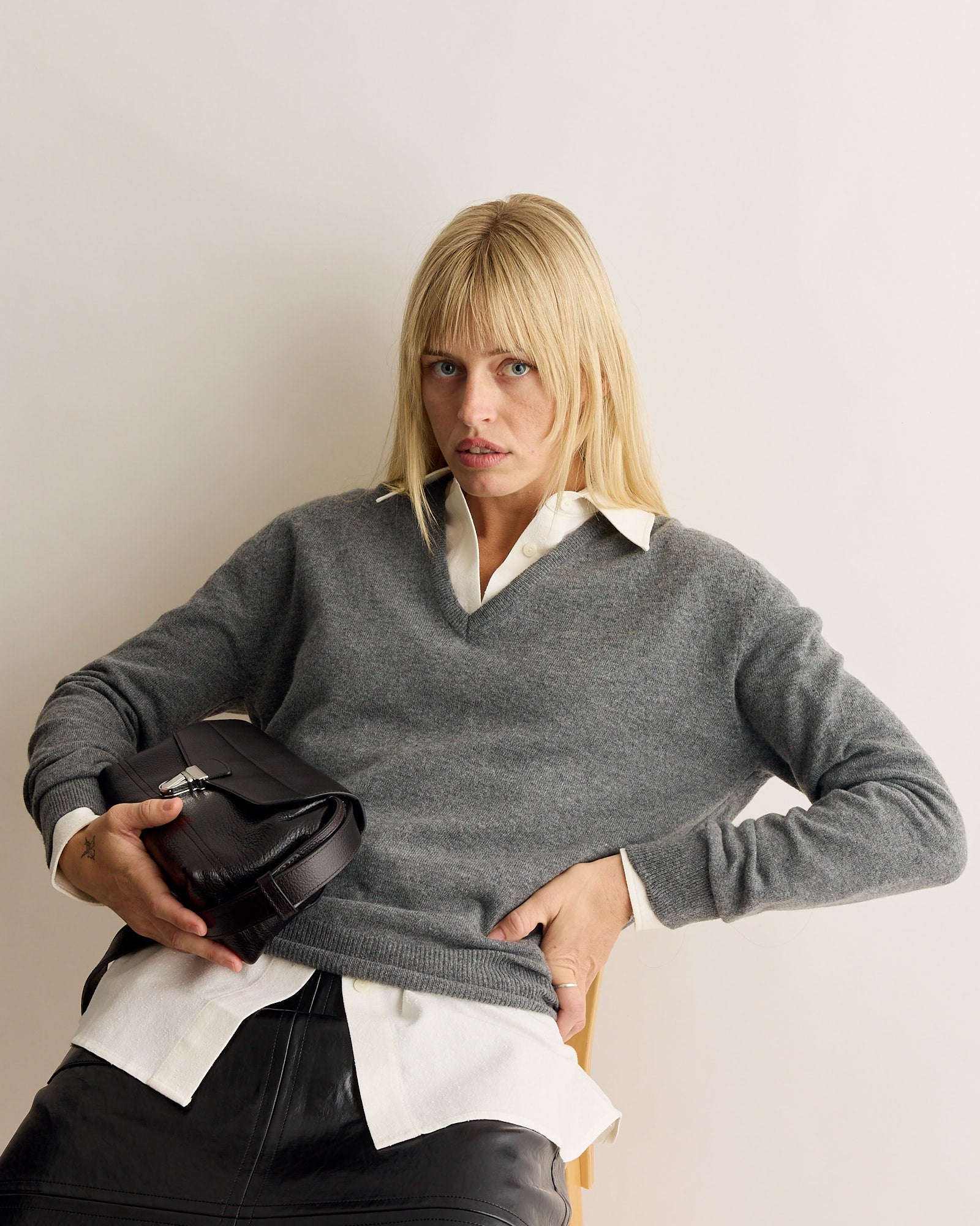 A person with straight blonde hair sits against a plain background, wearing the Mijeong Park Cashmere Blend V-Neck Knit in Grey over a white collared shirt and black pants, holding a black handbag on their lap.
