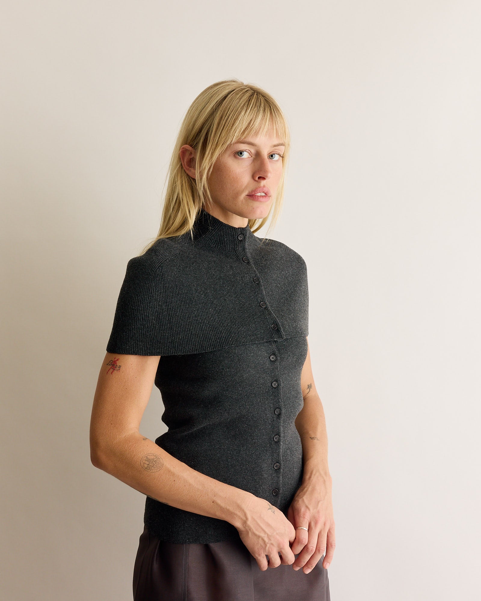 A person with straight blonde hair wears the Amomento Cape Top & Bandeau Set in Charcoal, featuring short sleeves, a high collar, and buttons, standing against a plain light background and gazing slightly to the side.