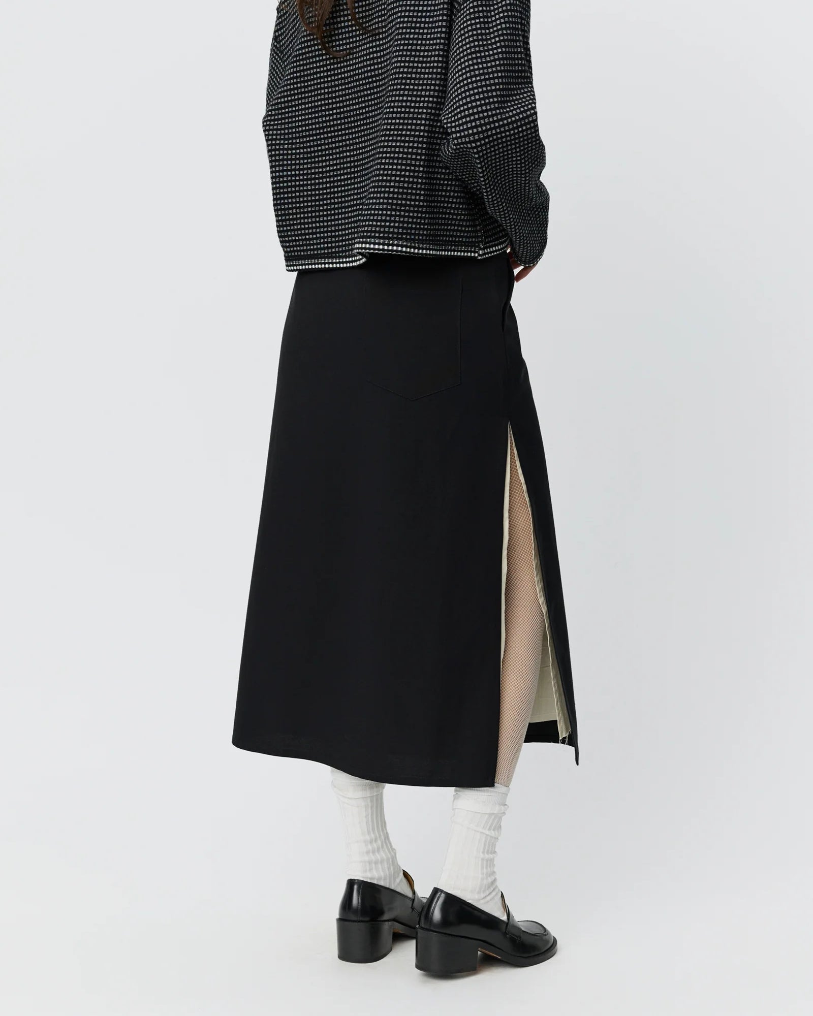 A person, visible only from the waist down, wears the mfpen Tab Skirt in Muted Black with a high side slit, a patterned black mfpen jacket, white socks, and black loafers.