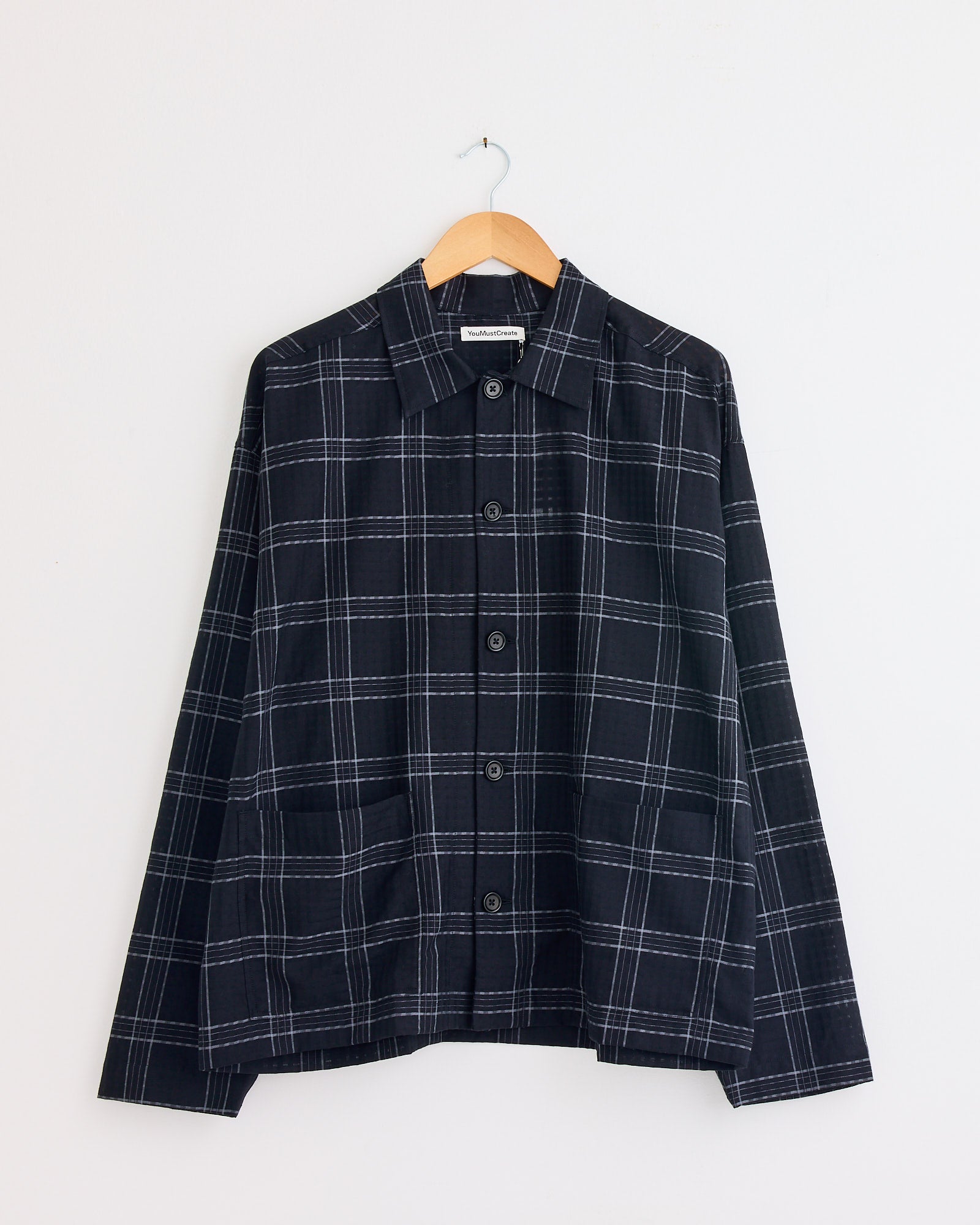 The YMC PJ Overshirt in Black/Grey features a white and gray plaid pattern, long sleeves, and a collar, displayed on a wooden hanger against a white wall. For fit or sizing questions, contact us via our online chat.