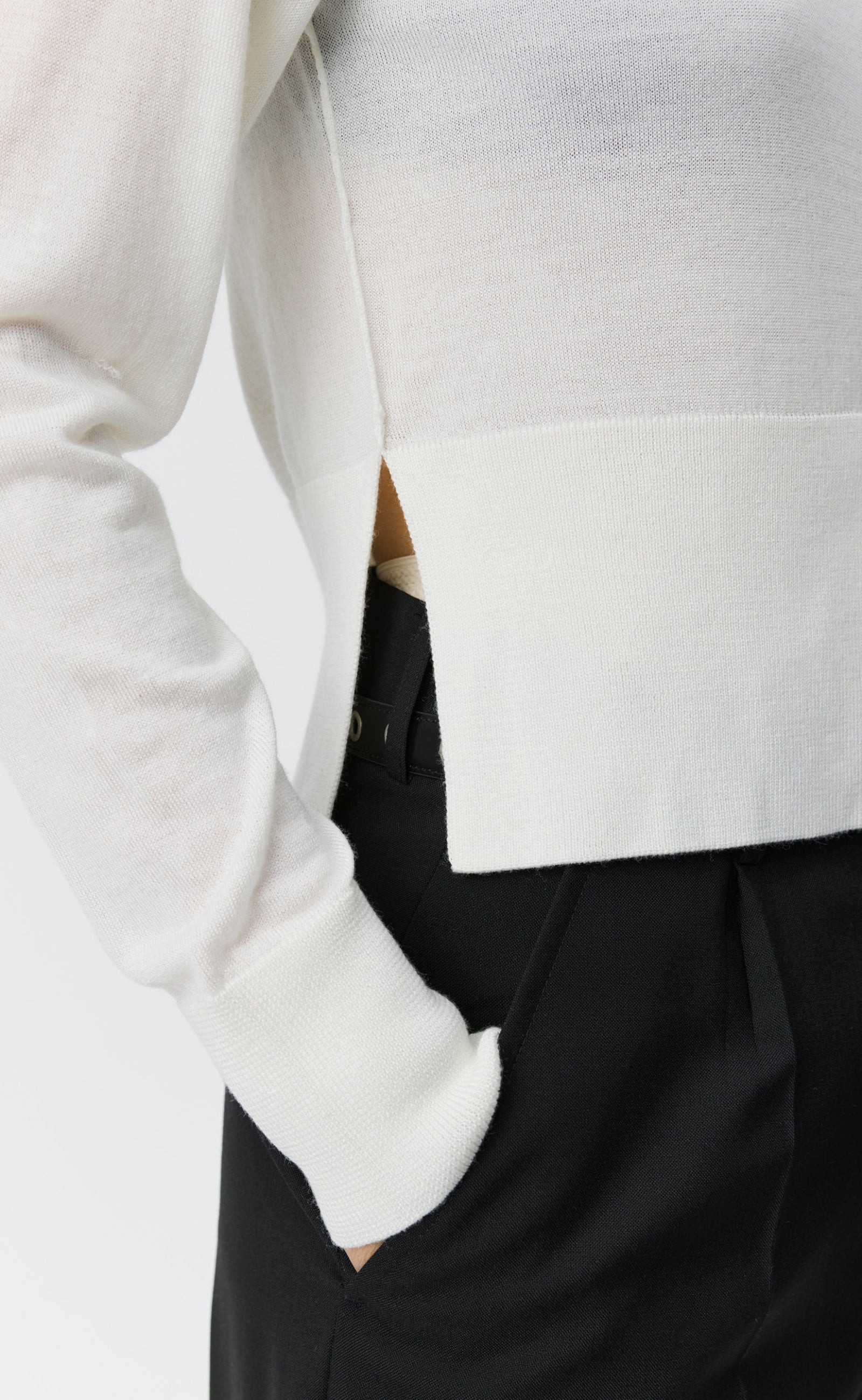 A person wears the mfpen Appointment Cardigan in Ecru with black trousers, standing with one hand in their pocket. The image is cropped to display only the torso and upper legs.
