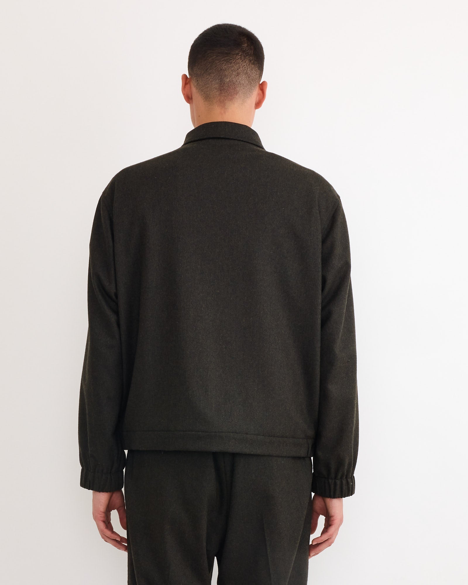 A person with short hair, facing away from the camera against a plain white background, is wearing the SMOCK Blakey Jacket in Cashmere Wool Dark Olive with matching pants.