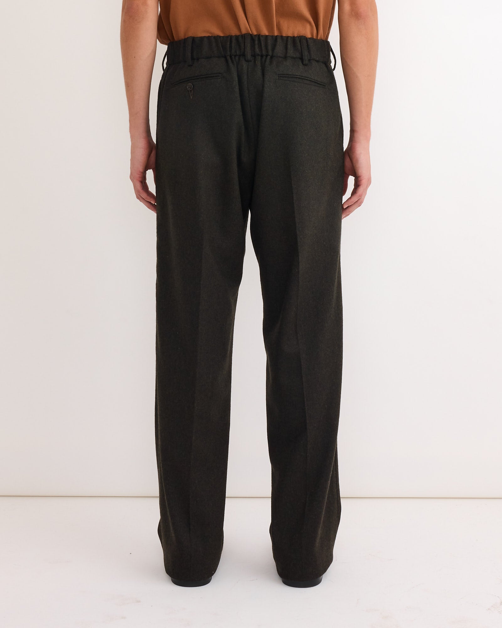 Person standing against a white background, wearing SMOCK’s West Coast Pant in Cashmere Wool Dark Olive with an elastic waistband and back pockets, paired with a brown top. The image is taken from behind to show the full pants length.