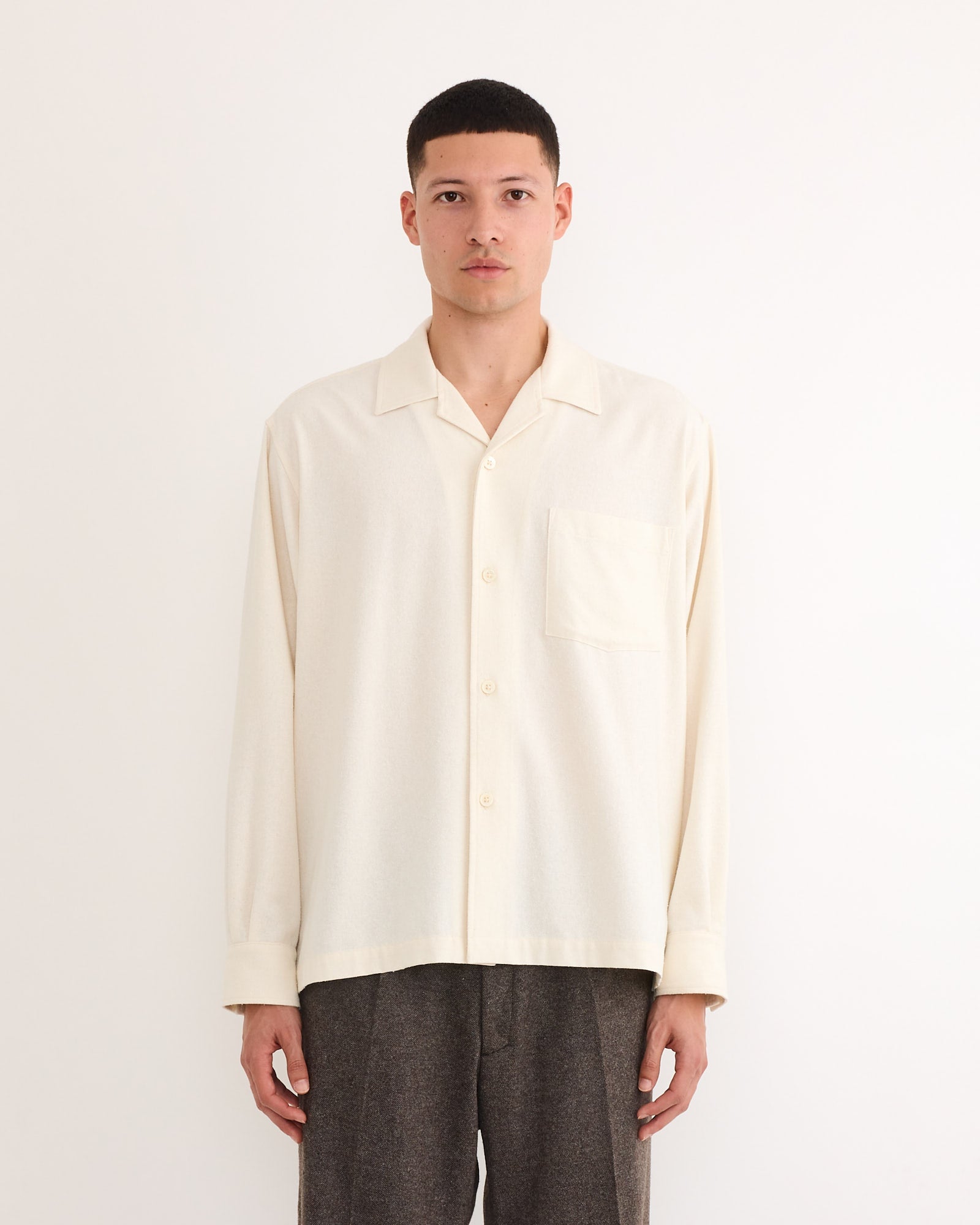 A person stands facing forward against a plain white background, wearing the SMOCK Evans Shirt in Raw Silk Natural with a chest pocket and loose dark gray pants, made in Los Angeles.
