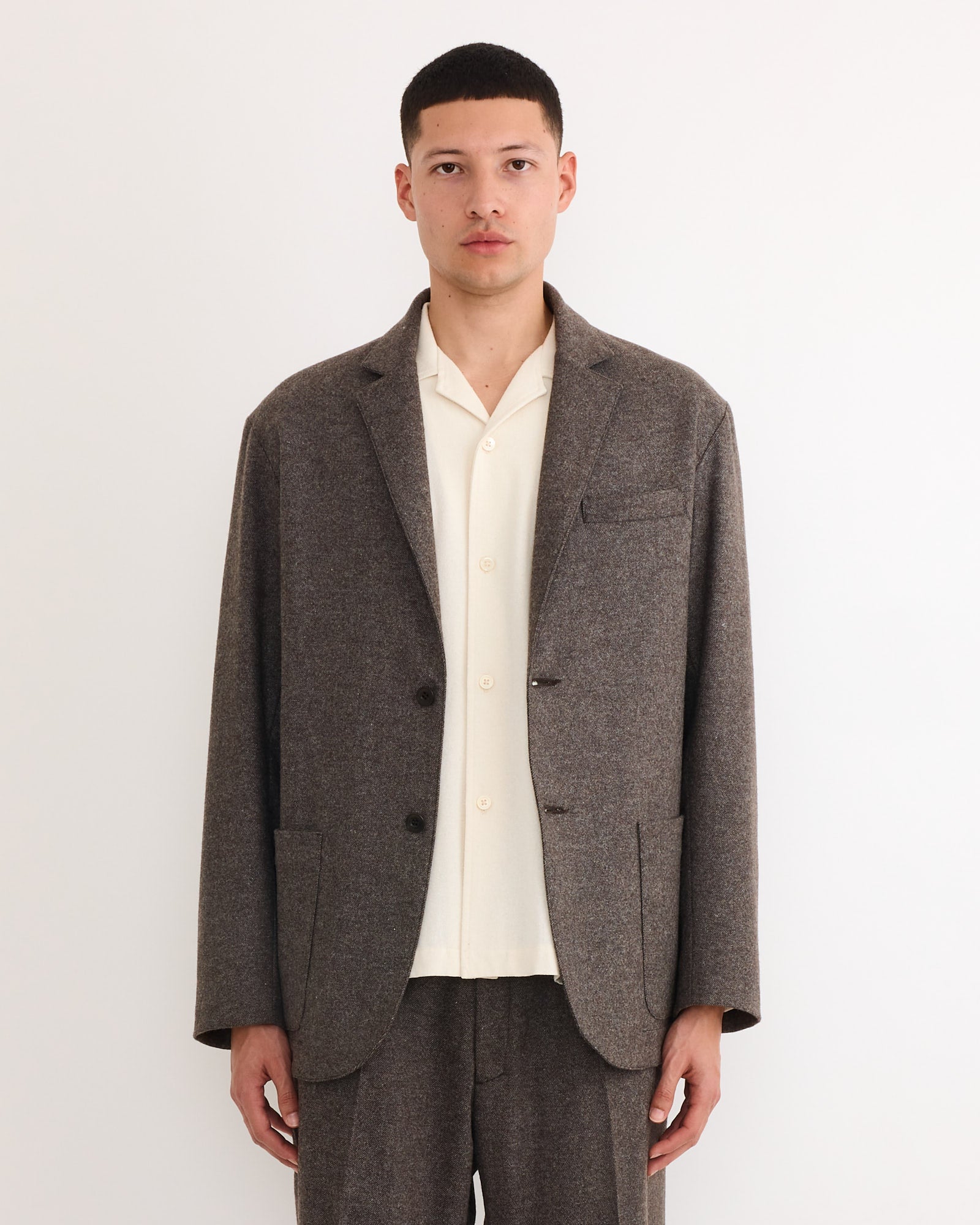 A person stands against a white background, wearing the SMOCK Blazer in Cashmere Wool Brown with matching trousers over a cream button-up shirt, looking directly at the camera with a neutral expression.