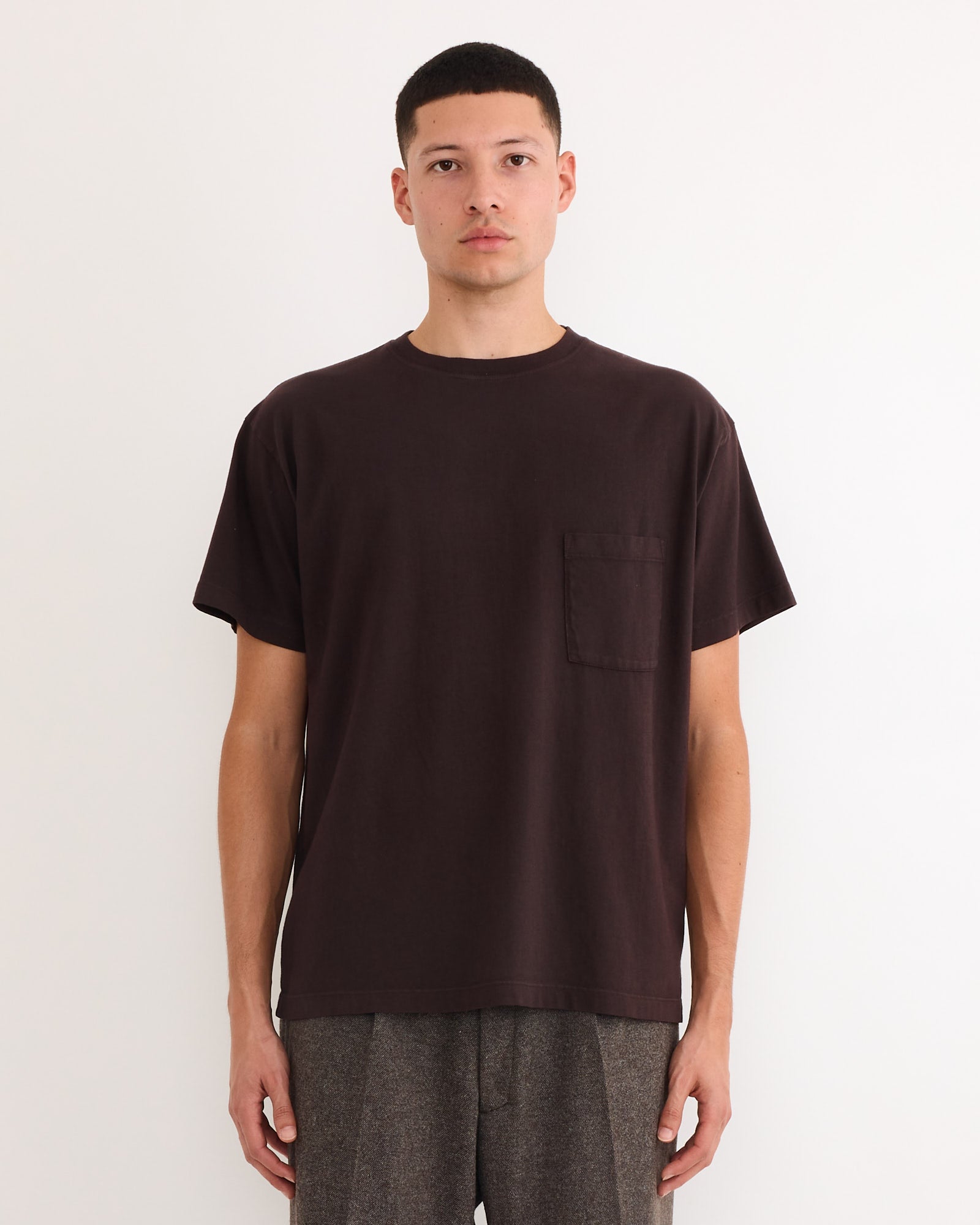 A person with short dark hair wears SMOCK’s Bachelor Pack T-shirt in Caper/Port/Chai with a chest pocket and gray pants, standing against a plain white background.