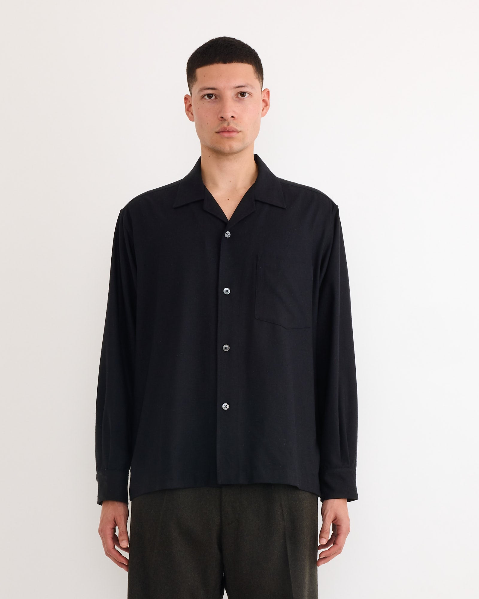 A person with short hair wears the SMOCK Evans Shirt in Raw Silk Black and dark pants, standing against a plain white background and facing forward with a neutral expression.