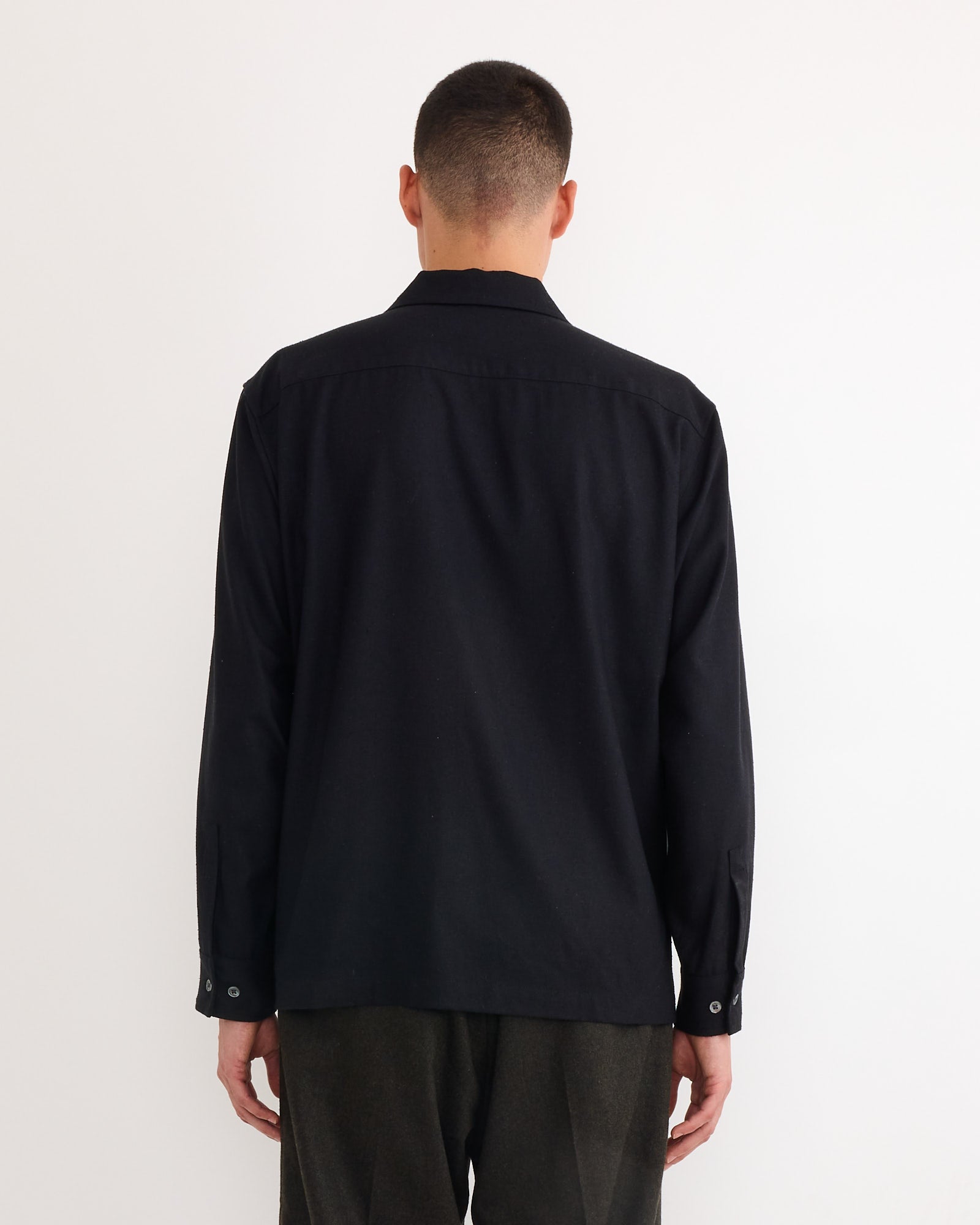 A person with short hair, facing away from the camera, wears the SMOCK Evans Shirt in Raw Silk Black and dark pants against a plain white background.