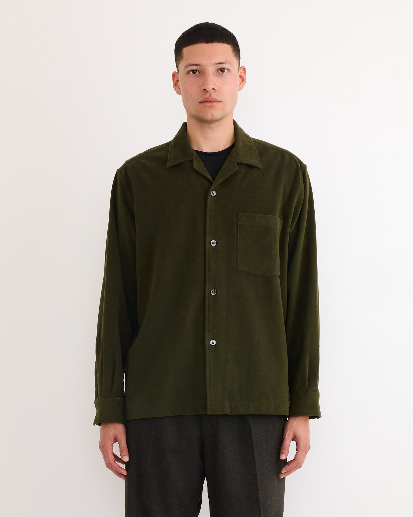 A person with short dark hair and a neutral expression stands against a plain white background, wearing the SMOCK Evans Shirt in Moleskin Green—an olive-green cotton long-sleeve shirt with chest pocket and dark trousers. Made in Los Angeles.