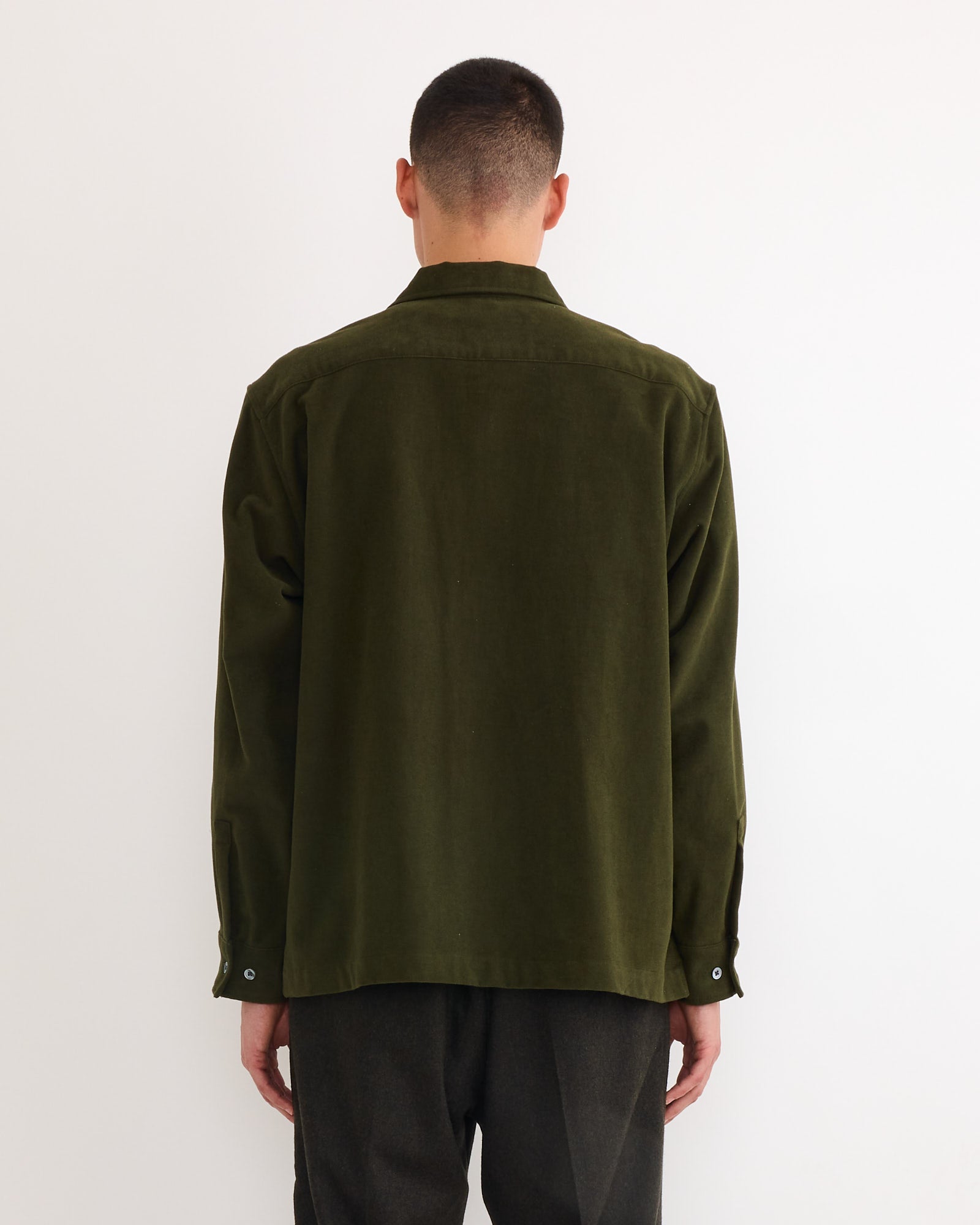 A person with short hair stands against a plain white background, facing away from the camera, wearing the SMOCK Evans Shirt in Moleskin Green and matching dark pants.