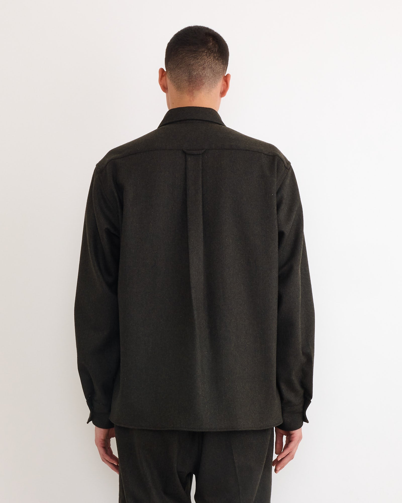 A person with short dark hair faces a white wall, wearing SMOCK's Davis Shirt in Cashmere Wool Dark Olive, made in Los Angeles and paired with matching pants. The back view showcases the outfit's simple style and quality fabric.