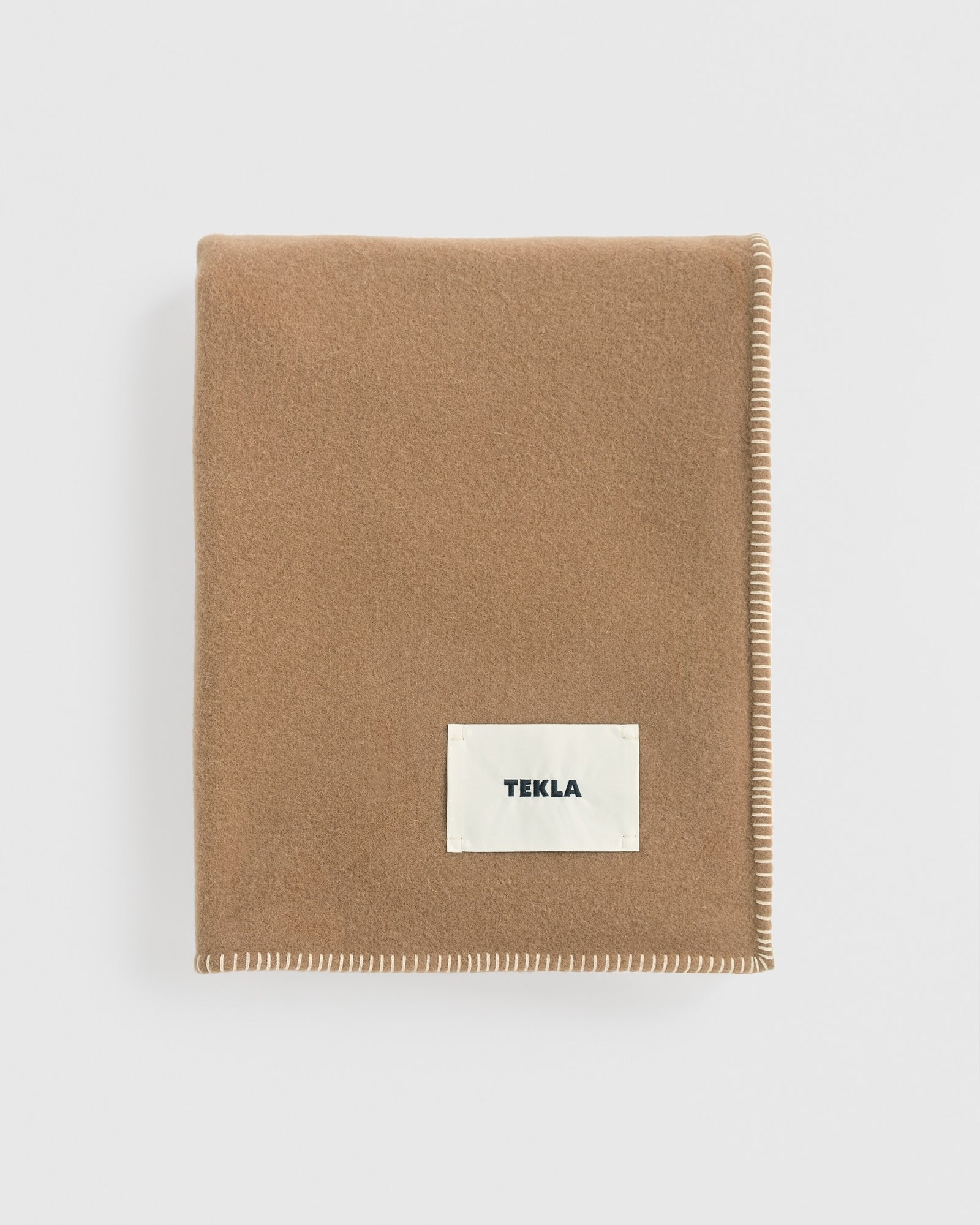 A folded TEKLA Lambswool Blanket in Oak with white stitched edges and a rectangular white TEKLA label, displayed against a plain white background.