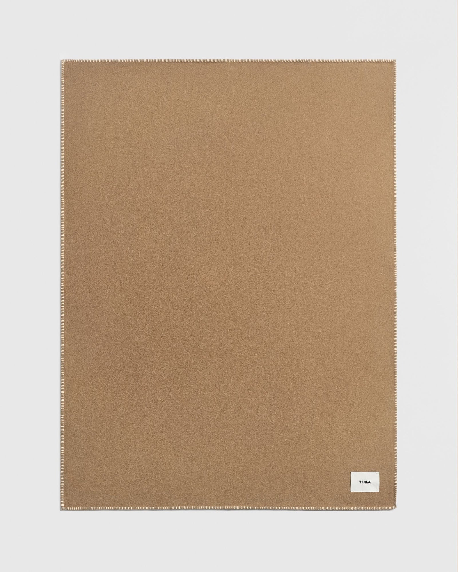 A plain, rectangular, light brown Lambswool Blanket in Oak from TEKLA with neatly stitched edges. A small white label with black text reading TEKLA is in one corner. The background is white.