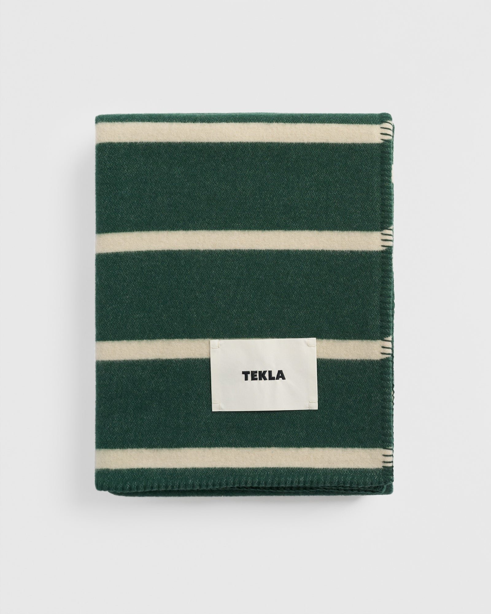 The TEKLA Lambswool Blanket in Rugby Stripes is a soft green lambswool blanket with wide cream horizontal stripes and a white TEKLA label, neatly folded and displayed on a plain white background.