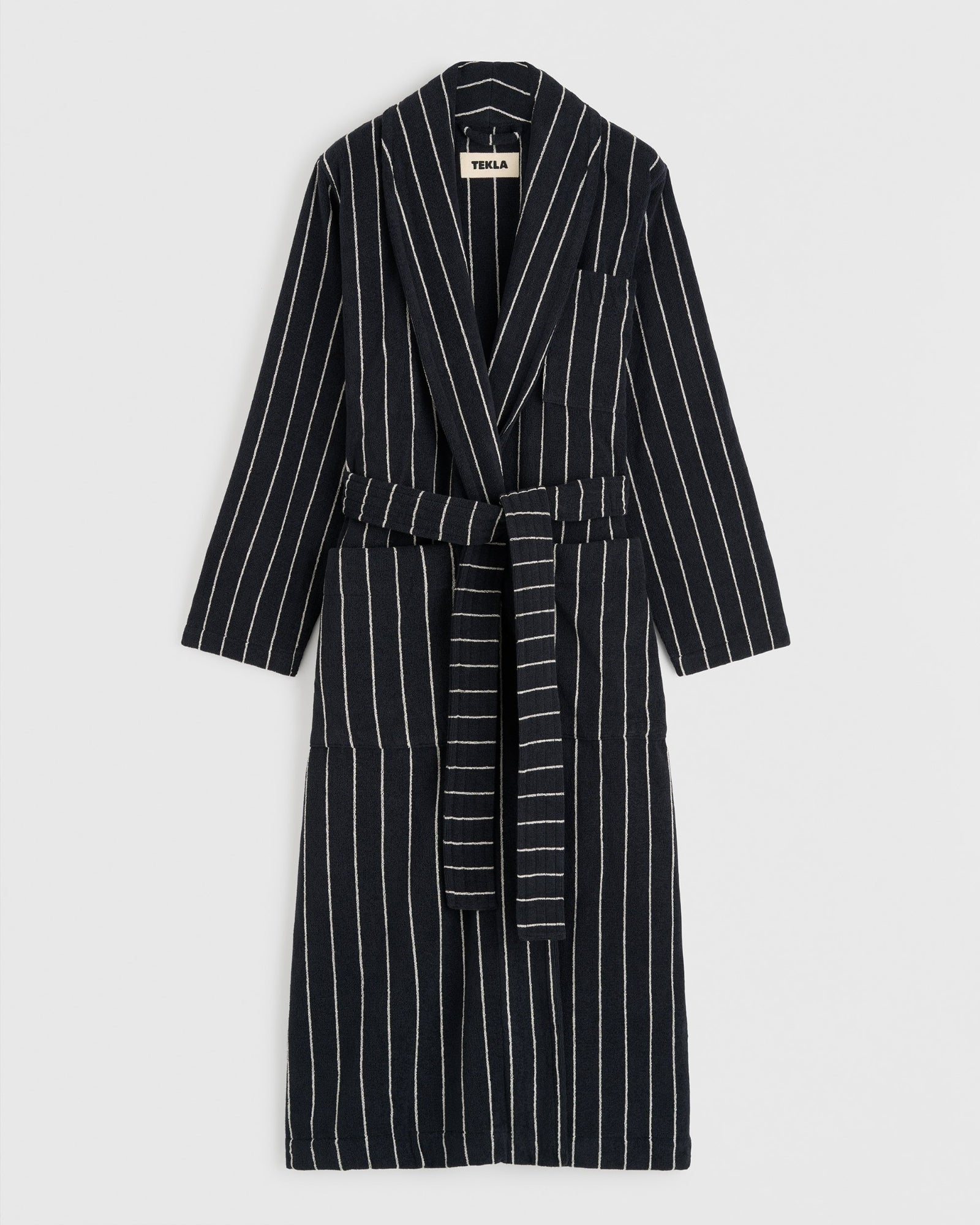 The TEKLA Classic Bathrobe in Antwerp is a long black organic cotton robe with white pinstripes, notched lapels, chest pocket, and a matching waist belt, shown flat on a white background.