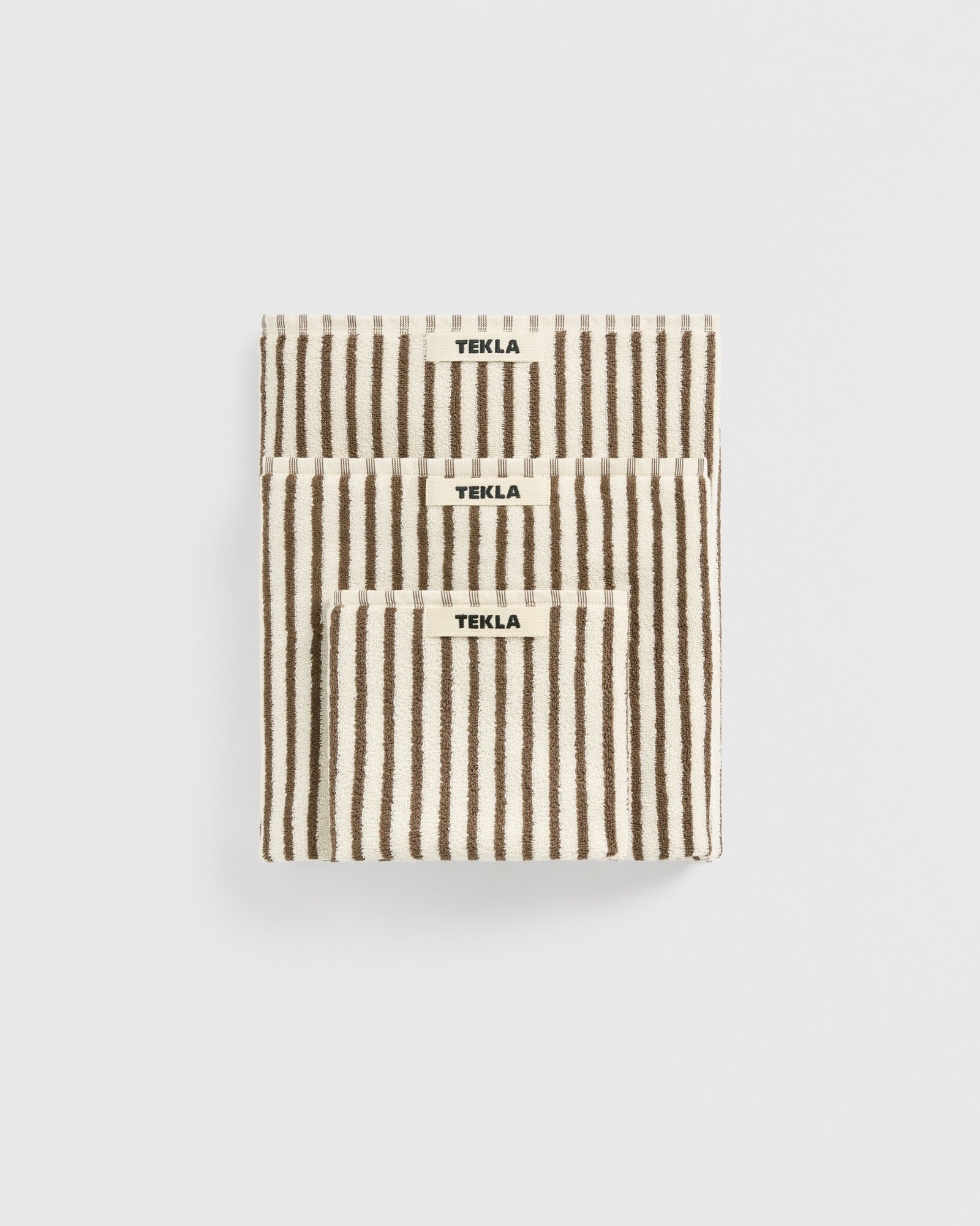 Three folded beige towels with vertical brown stripes, labeled “TEKLA,” are stacked neatly against a plain white background. The Towel Bundle in Kodiak Stripes by TEKLA is crafted from organic cotton in Portugal for style and sustainability.