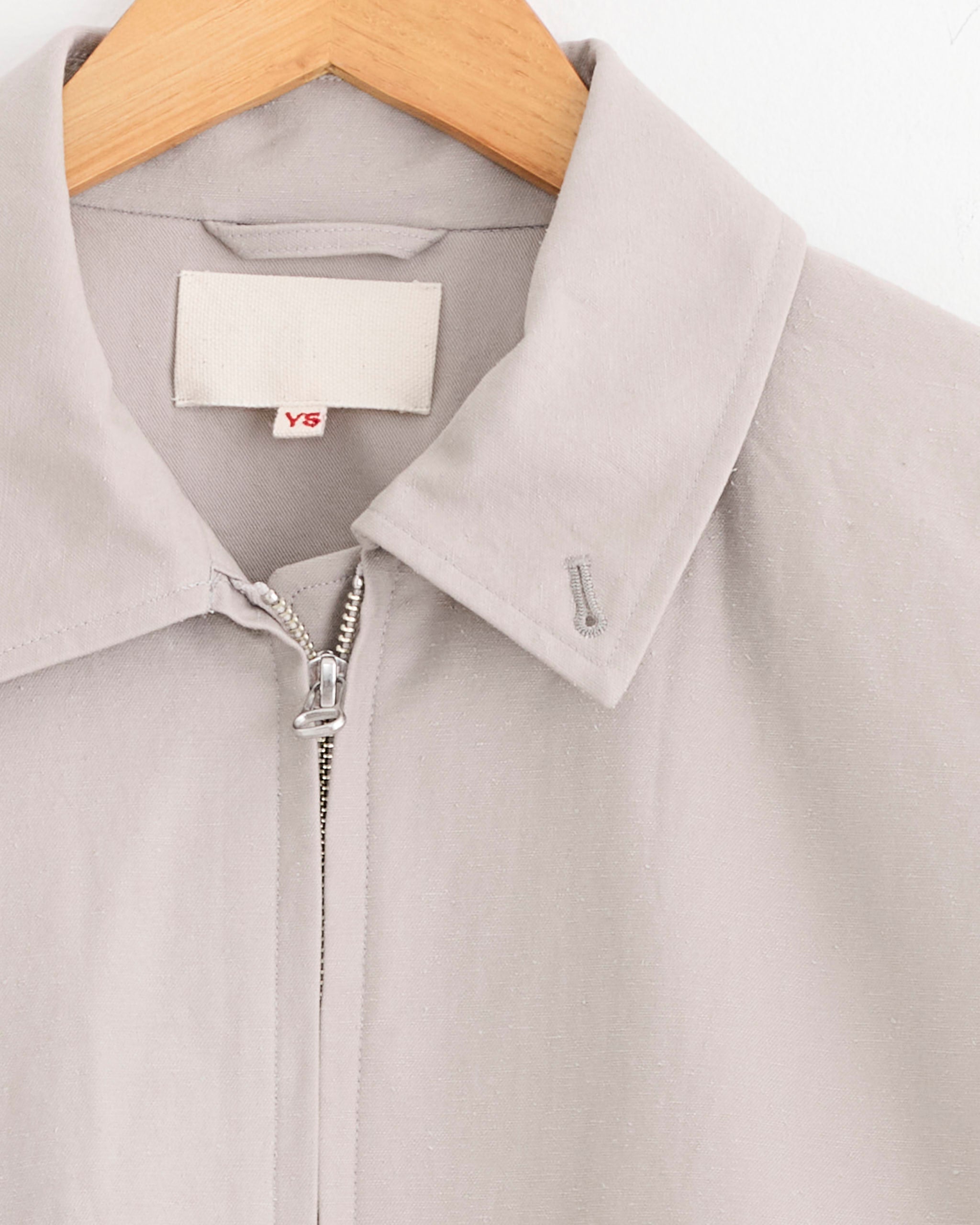 A close-up of the Yoko Sakamoto Zip Blouson in Beige, made from Japanese cotton, shown on a wooden hanger. Features a silver zipper, embroidered buttonhole, and a white YS label in red.