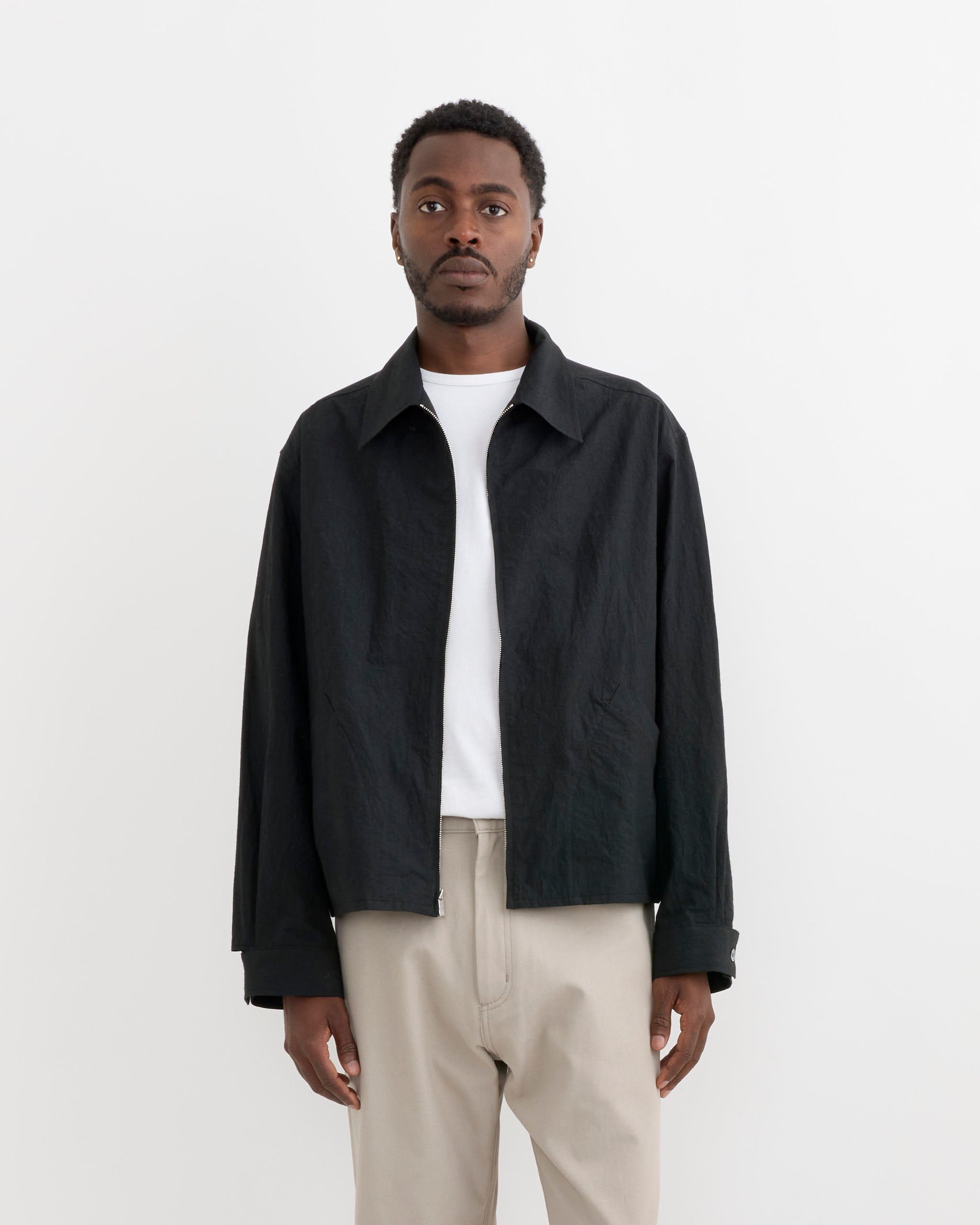 A man stands against a plain white background wearing the SMOCK 4H Jacket in Sun Dried Cotton Black over a white t-shirt and beige pants. Made from Japanese cotton typewriter fabric, he looks directly at the camera with a neutral expression.