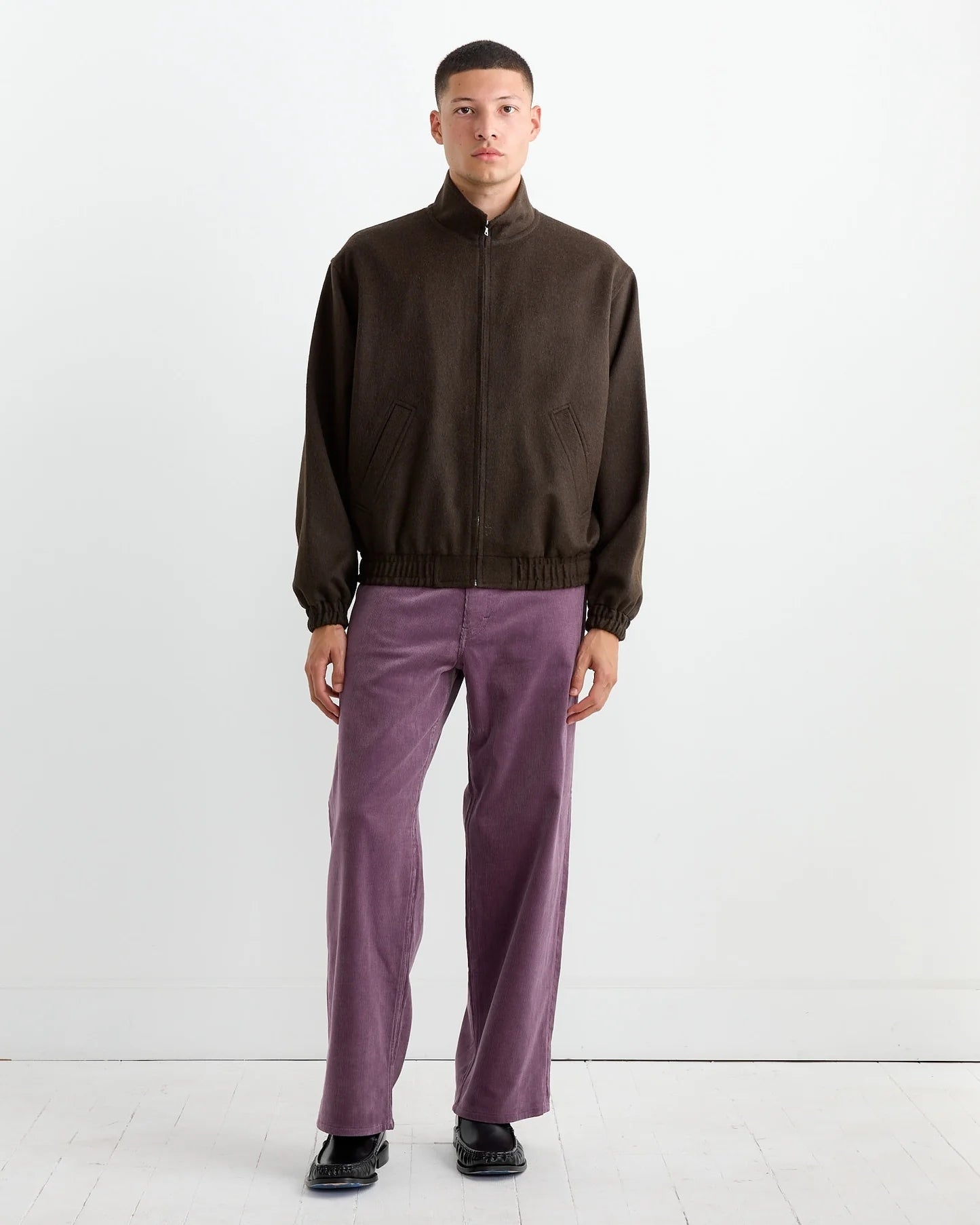 A person with short hair and a neutral expression stands against a white wall, wearing the Auralee Baby Camel Flannel Zip Blouson in Dark Brown, light purple pants, and black shoes. Their arms are relaxed at their sides.