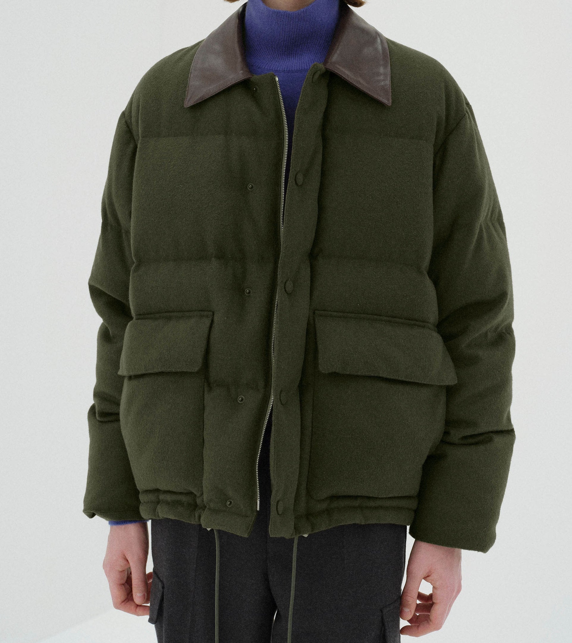 A person wearing the Auralee Baby Camel Flannel Down Blouson in Dark Olive, featuring a dark brown collar and large front pockets over a blue turtleneck. Only the torso and arms are shown.