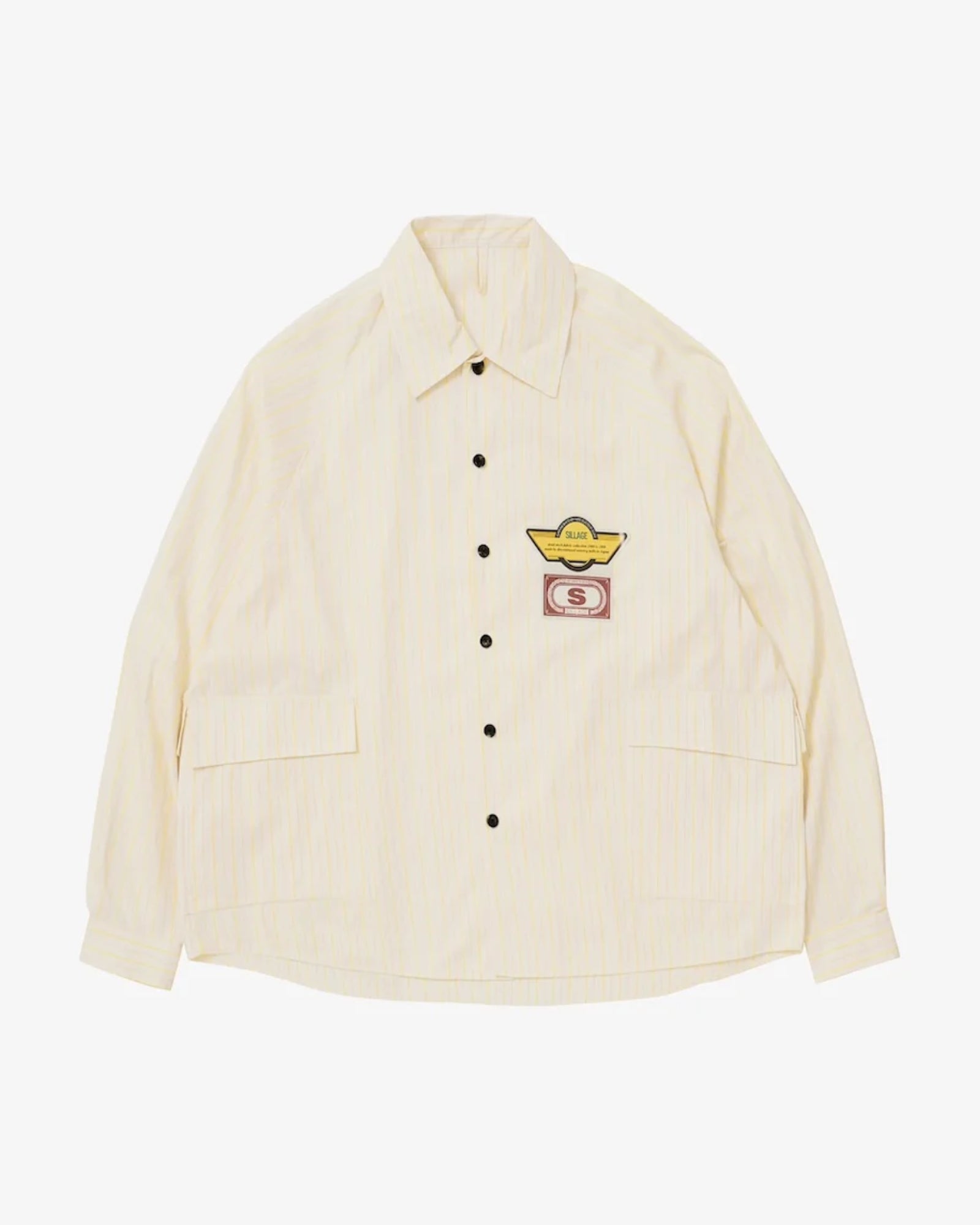 The Sillage Big Pocket Deadstock Overshirt is a cream, long-sleeve shirt with black buttons, two front pockets, and embroidered patches—a yellow winged design and a red “S”—on the left chest, crafted from vintage deadstock materials.