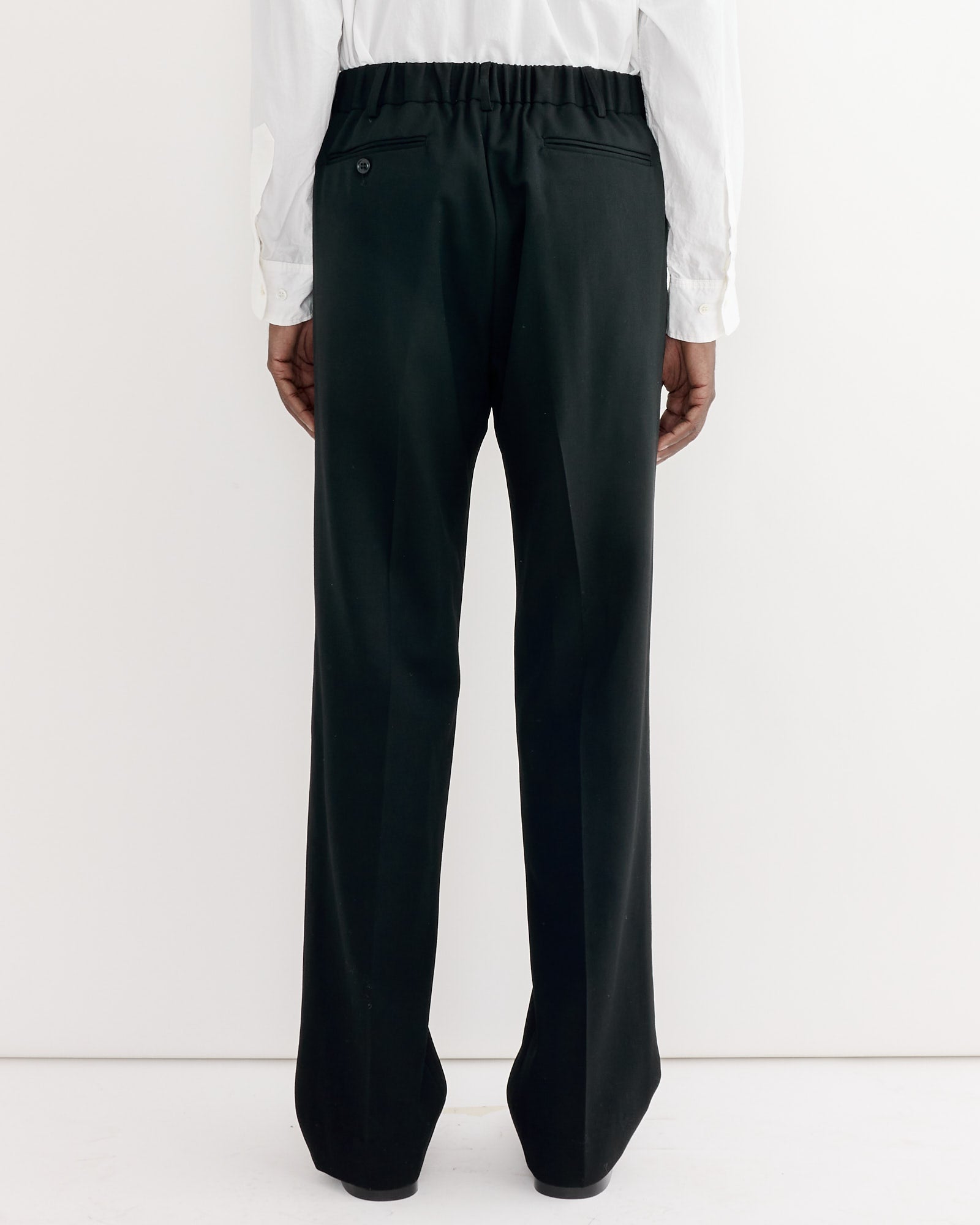 A person faces away from the camera, wearing SMOCK’s West Coast Pant in Wool Gabardine Black—men's pants made in Los Angeles with an elastic waistband, back pockets (one with a button), and a white long-sleeve shirt, against a white background.