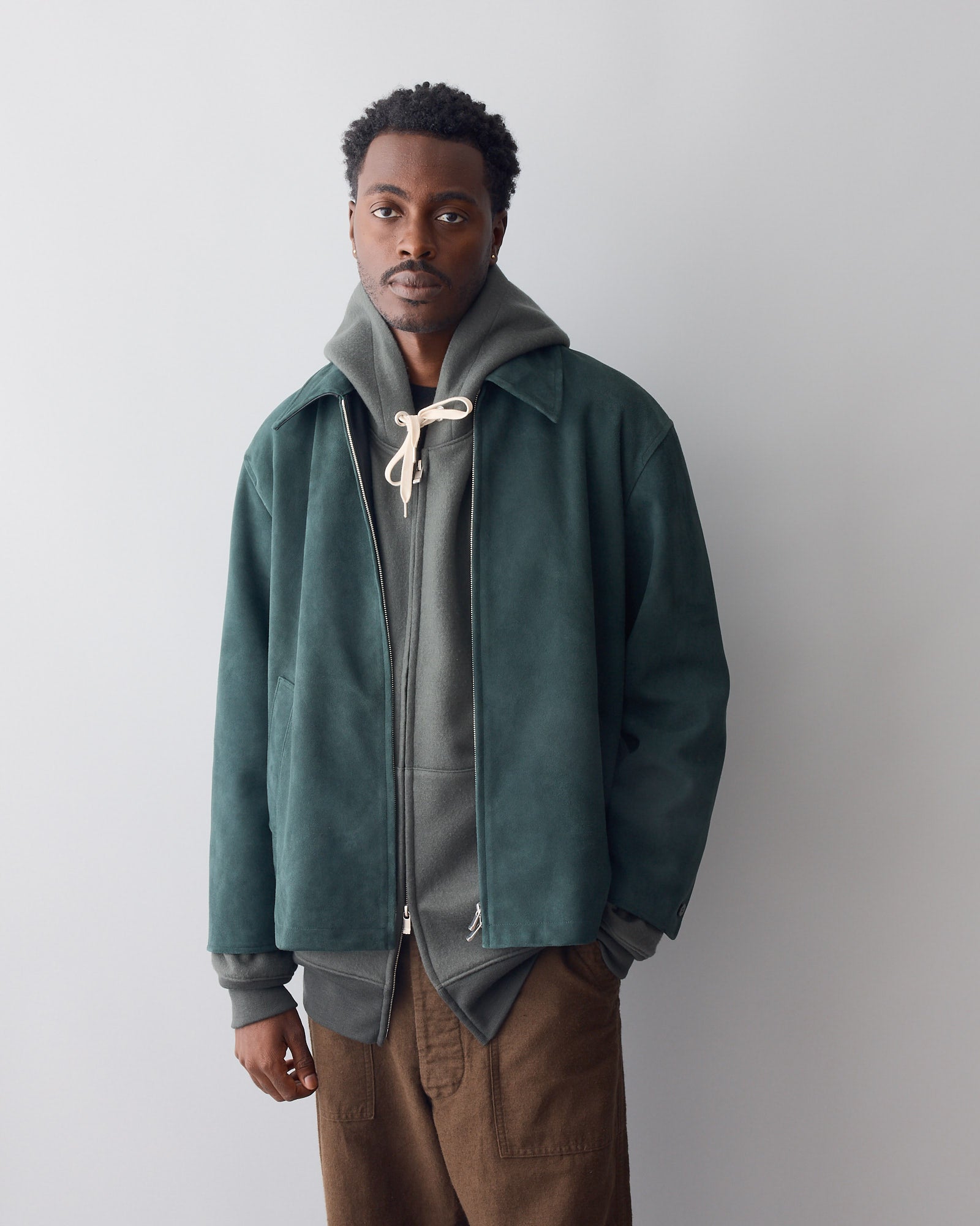 A man in the Cale Sheep Suede Heavy Jacket in Green stands before a plain light gray background, layered over a gray hoodie with cream drawstrings and brown pants, looking directly at the camera with a neutral expression.