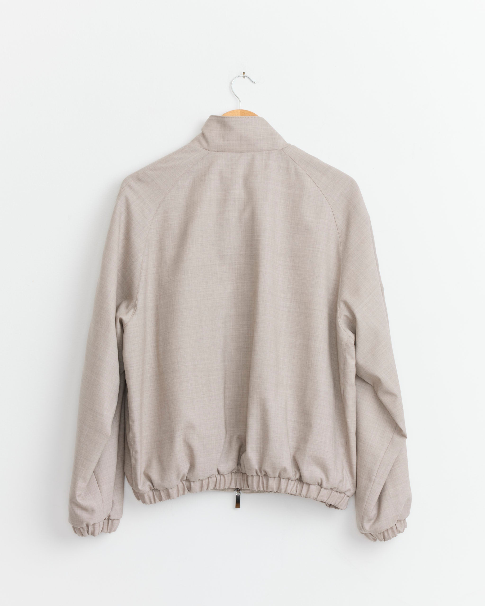 The Stoffa Reversible Blouson in Taupe, featuring long sleeves, elastic cuffs, and a gathered waistband, is displayed from the back on a wooden hanger against a white wall.
