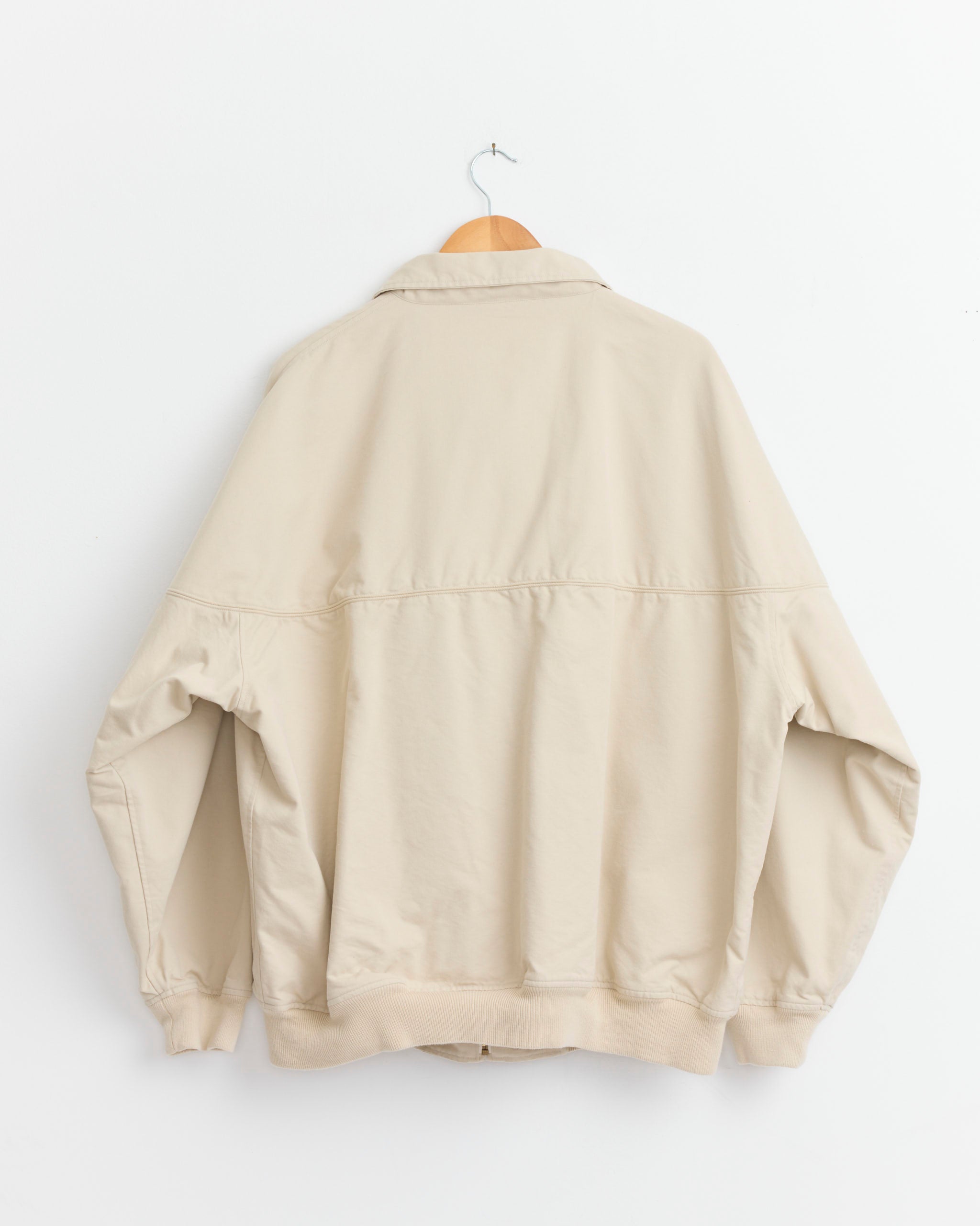 The Comoli Post Work Twill Cap Shoulder Blouson in Ivory hangs on a wooden hanger against a plain white wall, displayed with the back facing forward.