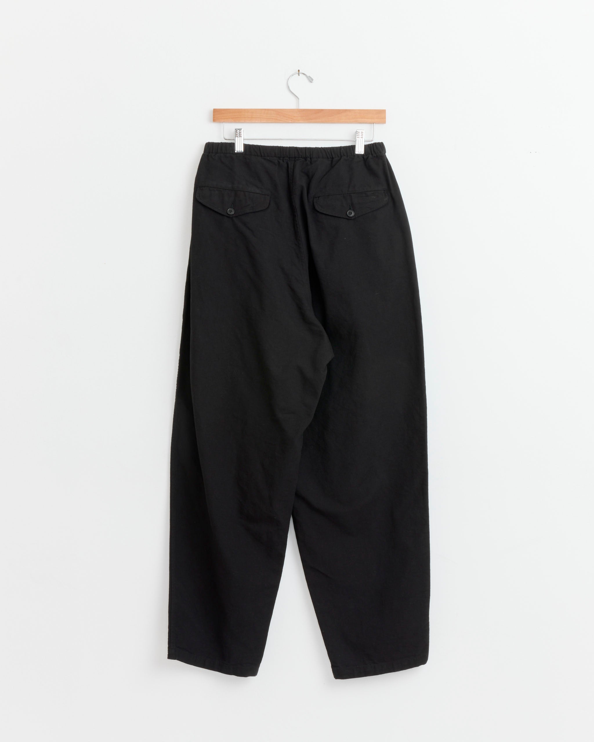 The Comoli Cotton Duck Tapered Pant in Black, featuring an elastic waistband and two buttoned back pockets, hangs on a wooden hanger against a plain white background.