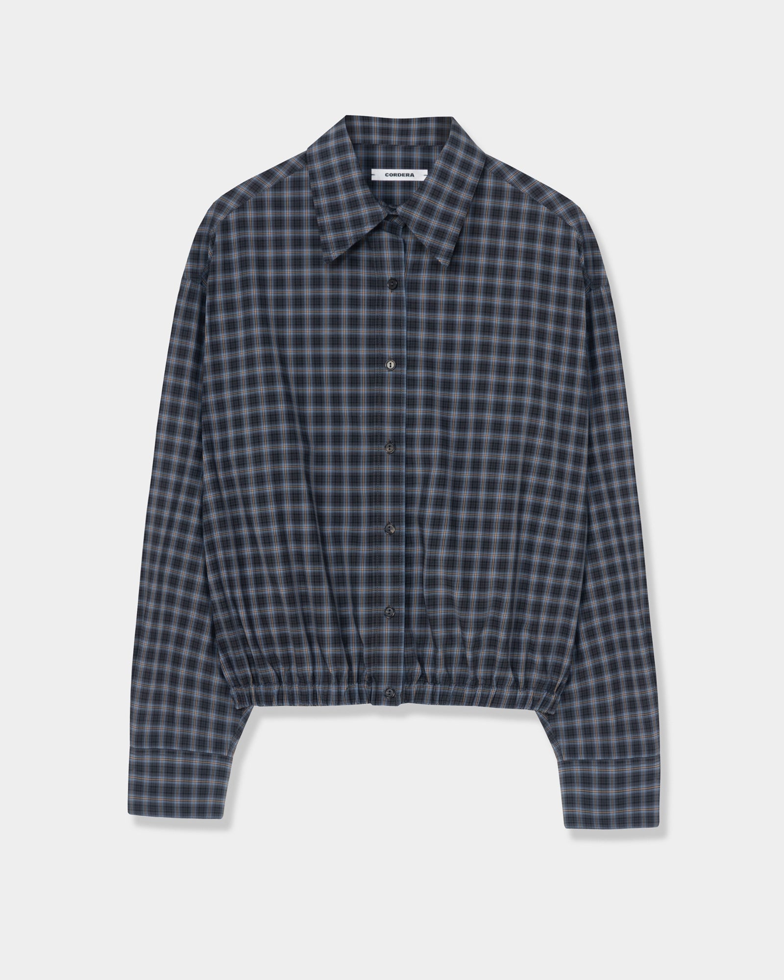 The Checked Bomber Shirt in Navy/Orange by CORDERA is an oversized, long-sleeve cotton shirt with a classic collar and elastic waistband for a subtle balloon silhouette, showcasing a navy and orange check pattern against a plain white background.
