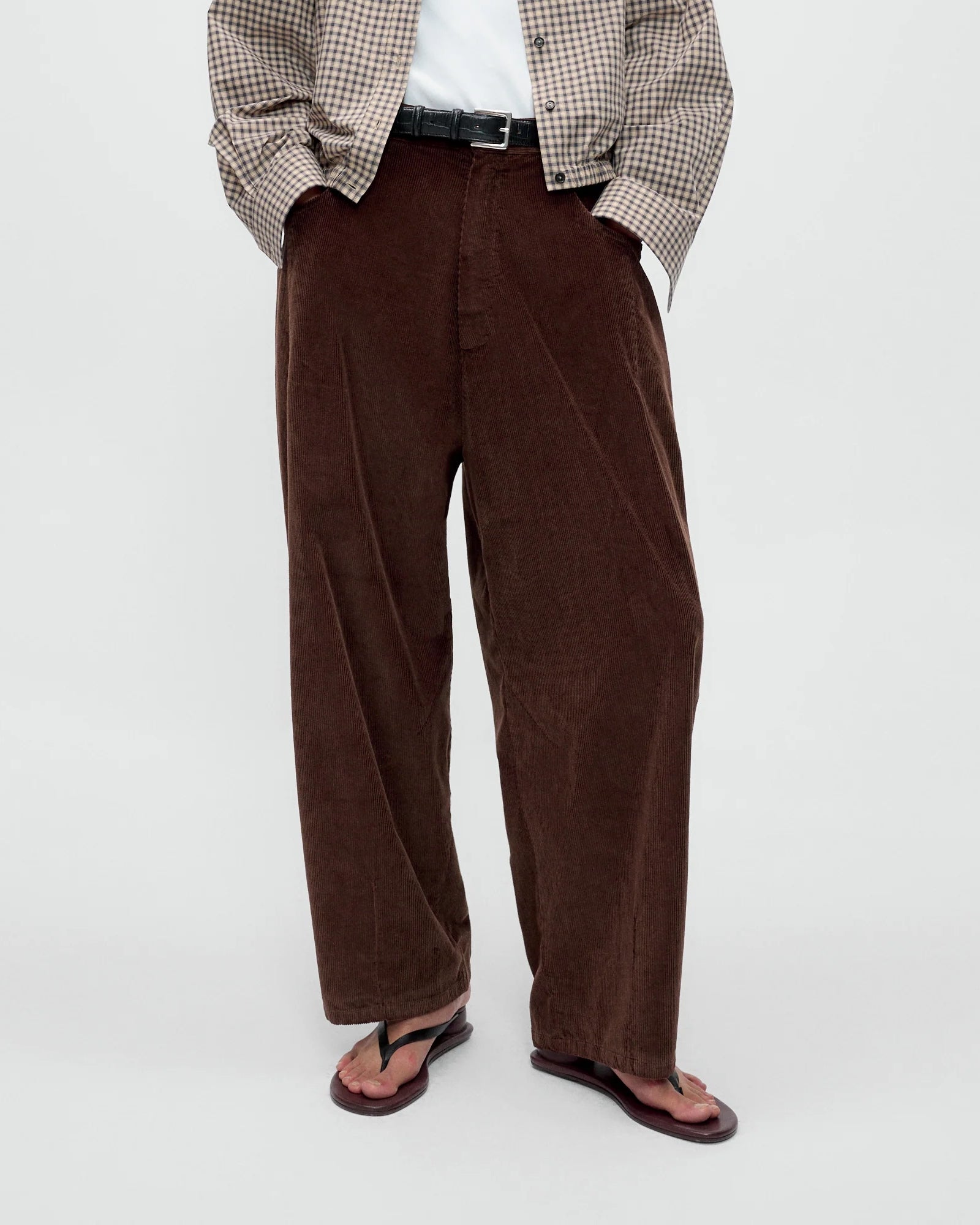 A person wearing Cordera’s Baggy Pant in Brown, paired with a white shirt, a checked jacket, and black flip-flops stands with hands in pockets against a plain background.
