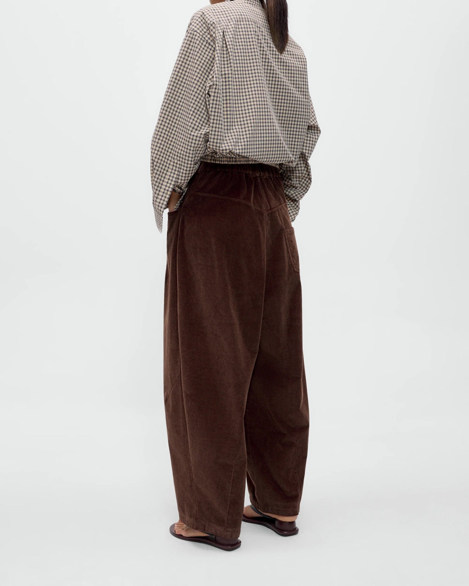 A person stands against a plain background, wearing the Cordera Baggy Pant in Brown paired with a checked long-sleeve shirt tucked in and black sandals. The image is shot from behind, with their head turned slightly.