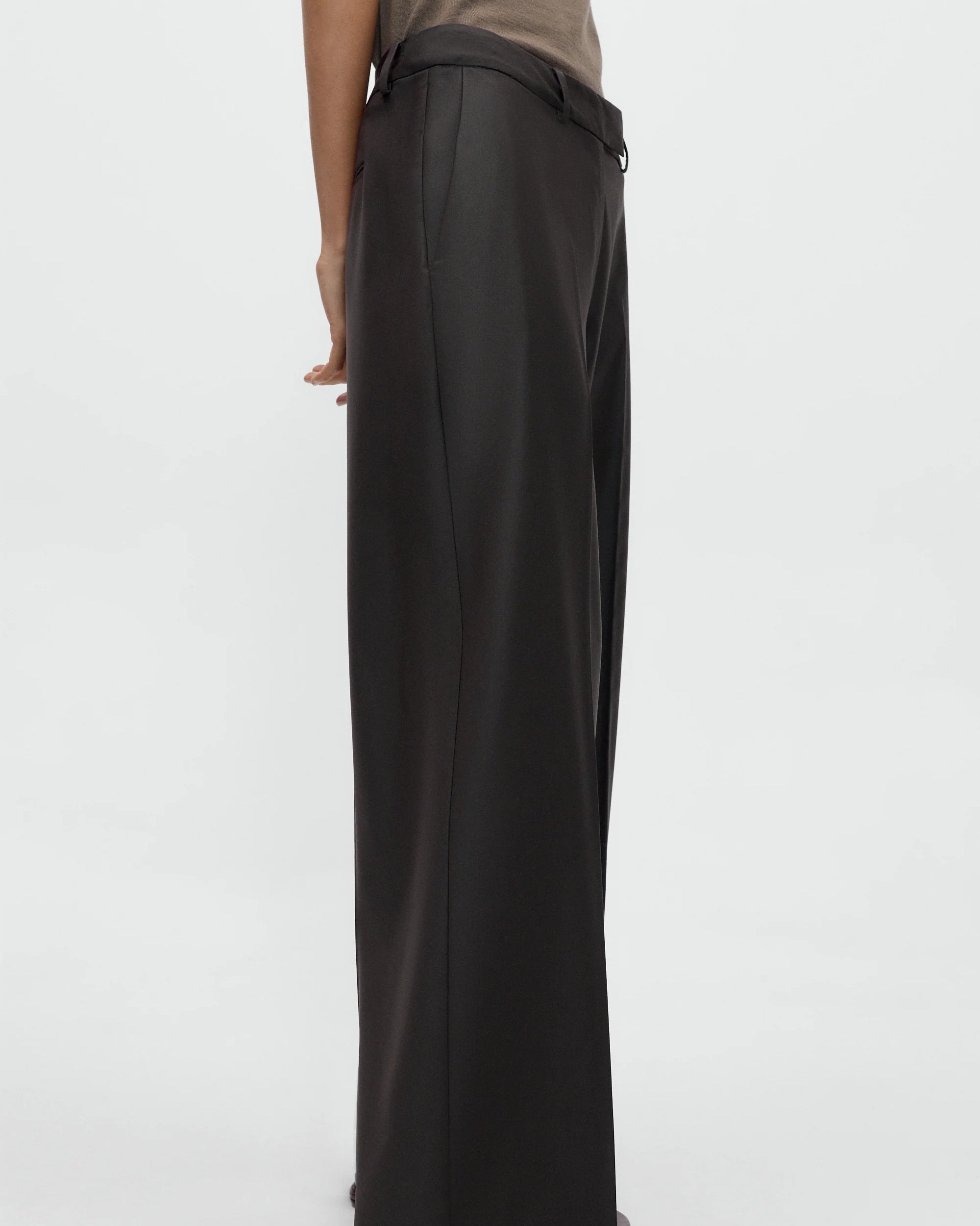 A person wearing CORDERA’s Low Rise Virgin Wool Pant in Dark Brown, styled with a tucked-in beige top, stands against a plain white background. Only the lower body and one arm are visible.