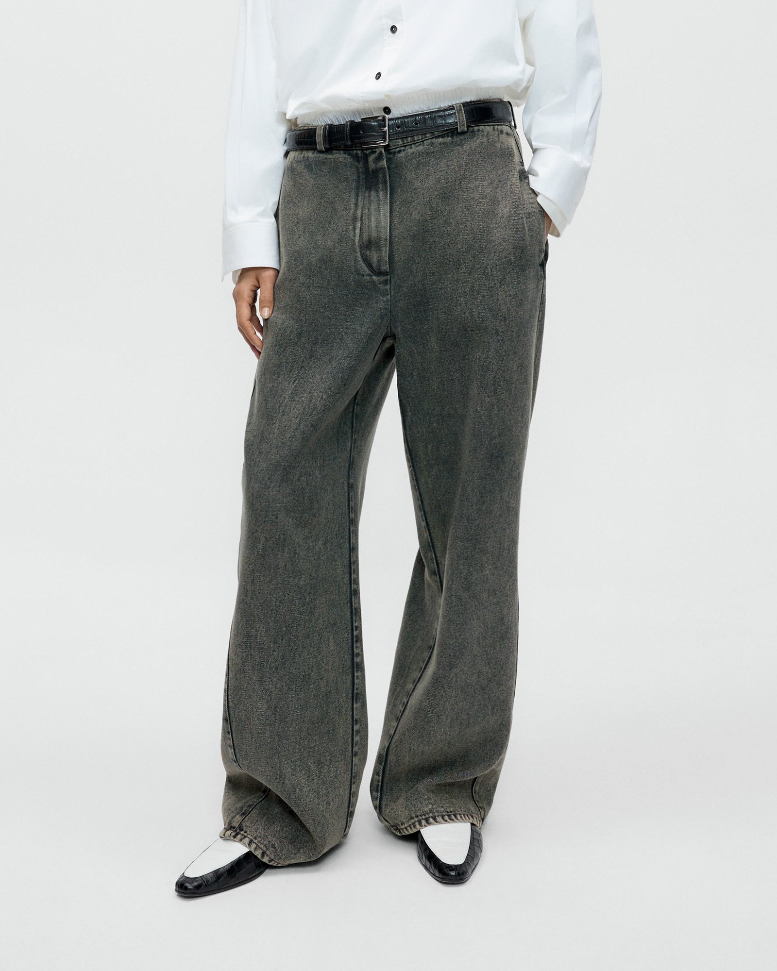 A person wearing a white button-up shirt, black belt, Cordera Washed Denim Straight Pant in Grey, and black and white pointed shoes stands against a plain white background.