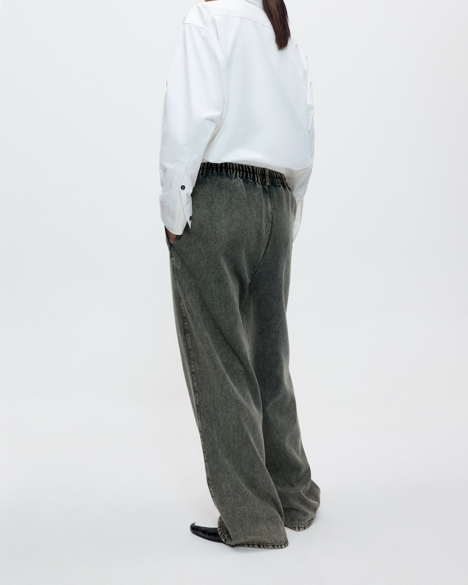 A person stands with their back to the camera against a plain white background, wearing loose-fitting Cordera Washed Denim Straight Pant in Grey and a white long-sleeve shirt.