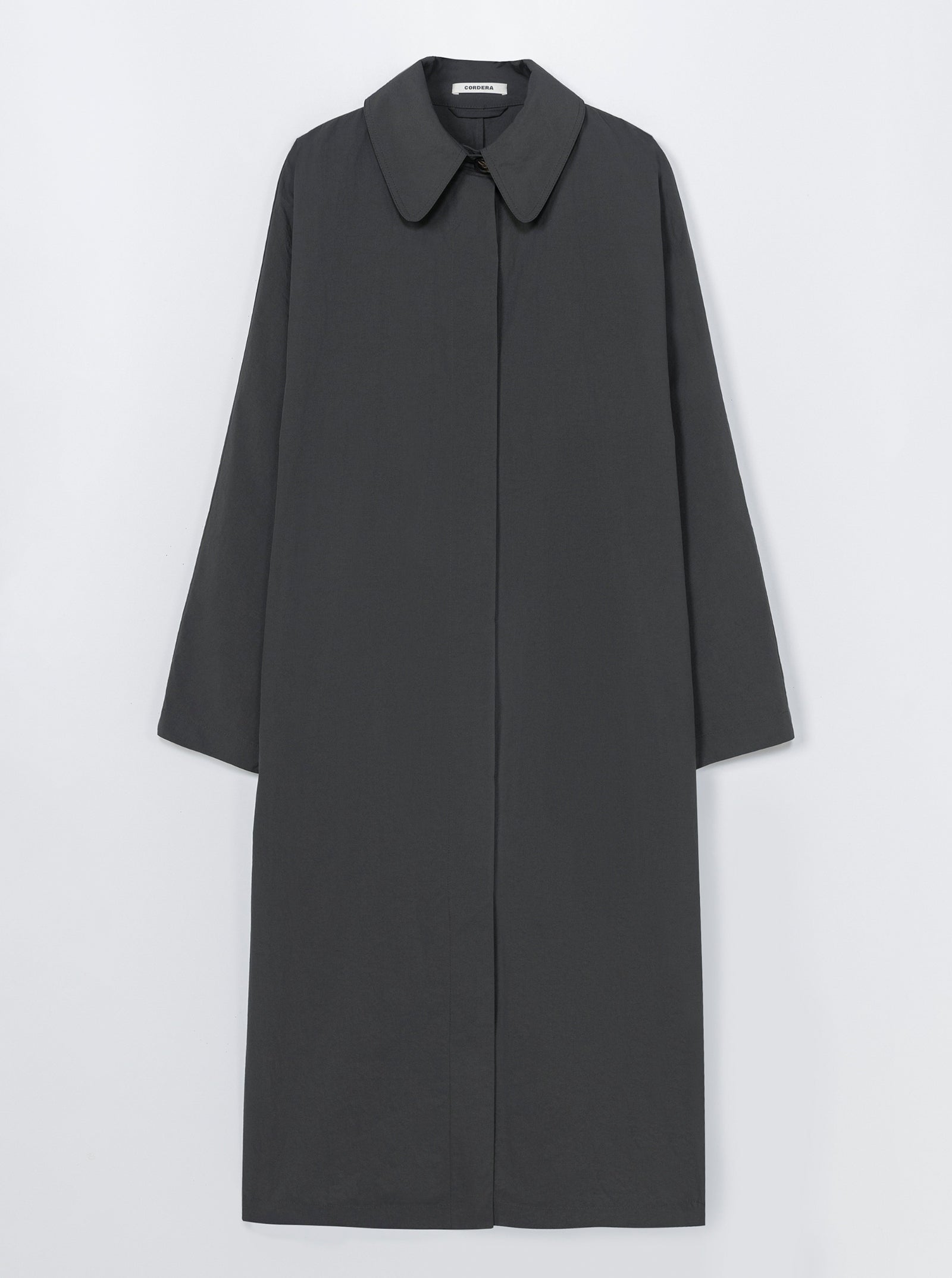 The Batwing Trench Coat in Ebony by CORDERA features a minimalist, dark gray design with a pointed collar and concealed buttons. Displayed on a plain white background. For sizing help, please use our online chat.