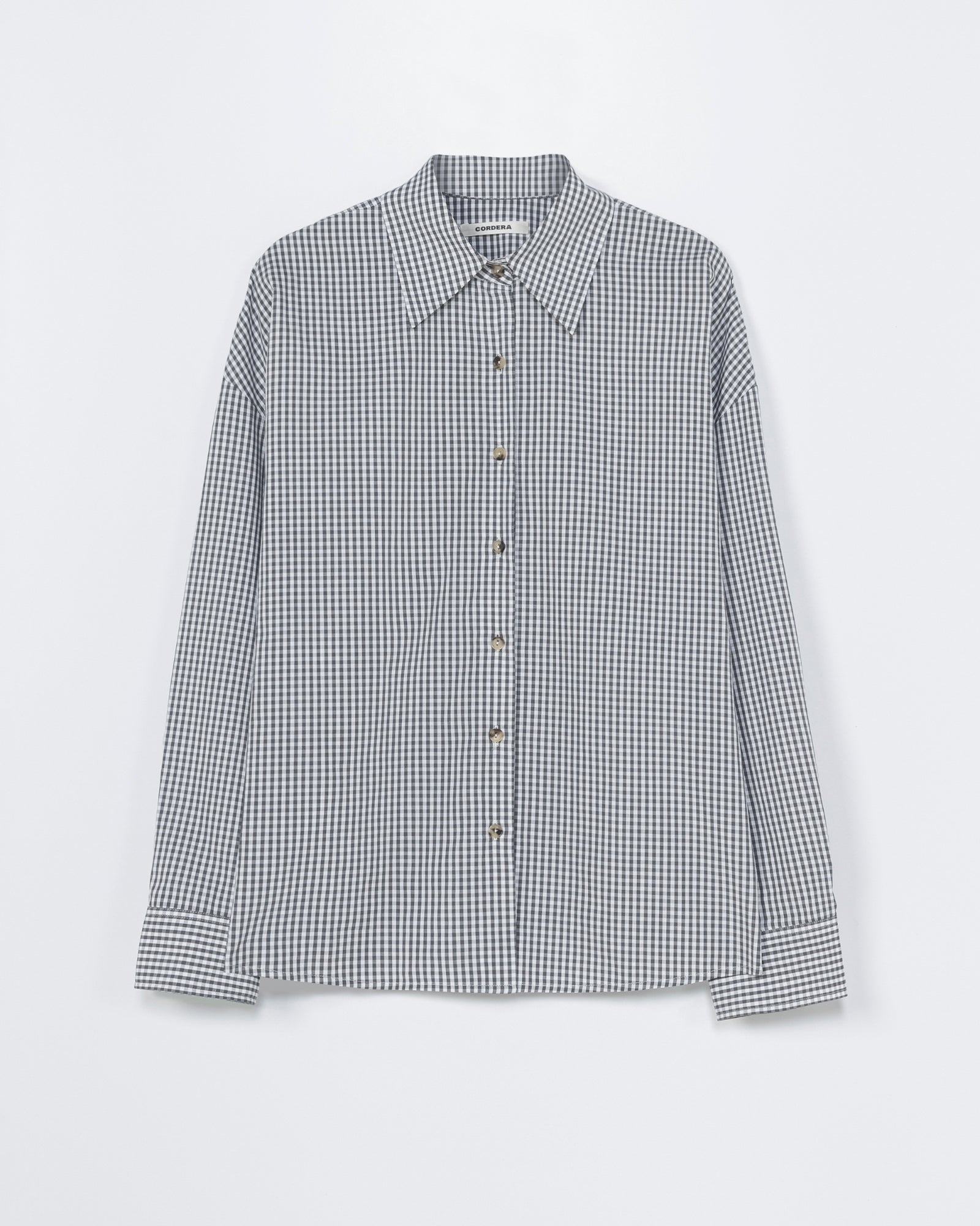 The CORDERA Oversized Shirt in Vichy Grey features long sleeves, a button-up front, pointed collar and cuffs, with a small black and white gingham check pattern. Displayed on a plain white background. For sizing help, chat with us online.