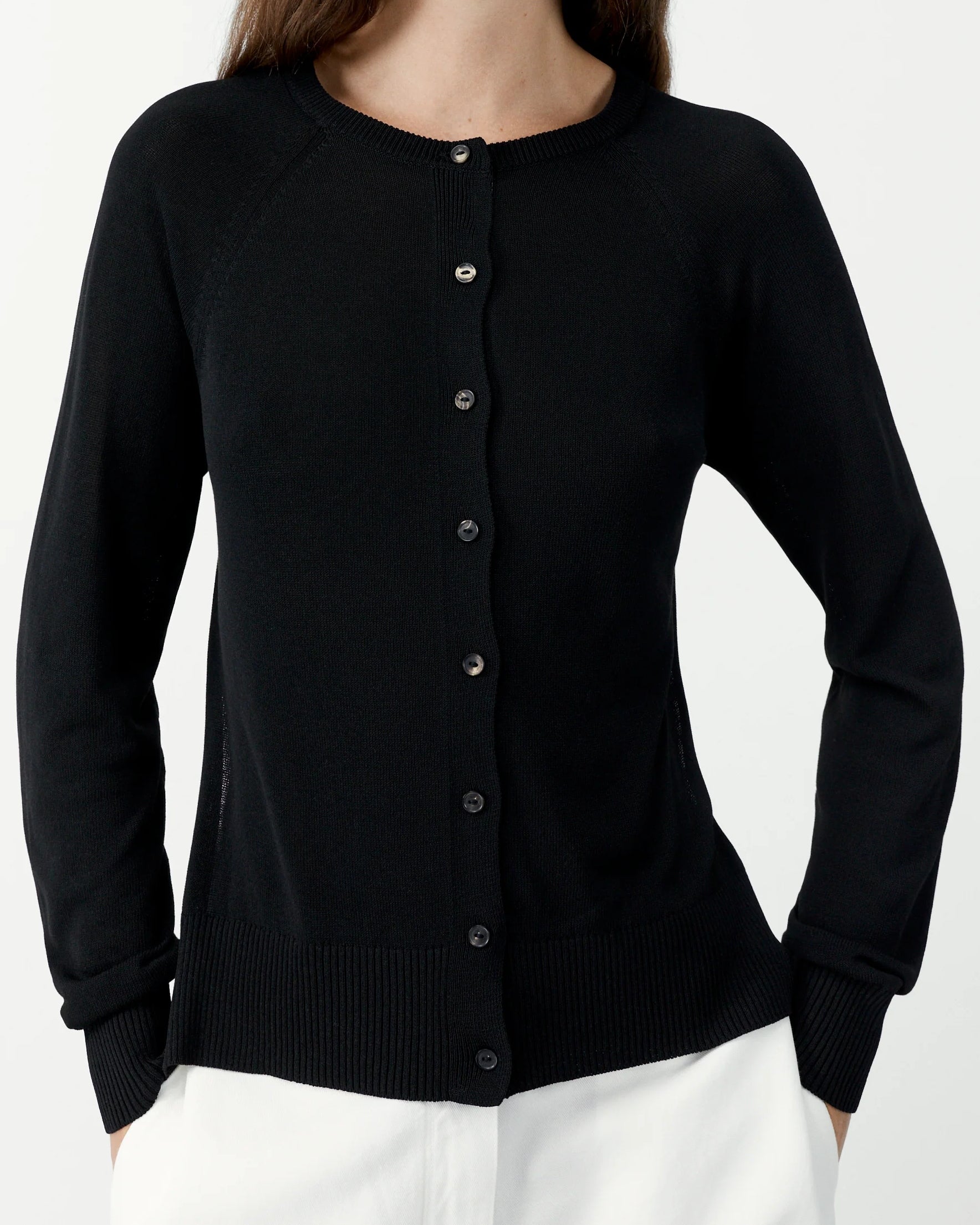 A woman wears the CORDERA Open Back Cardigan in Black—a lightweight, long-sleeve button-up knit with a round neckline—paired with white pants and standing against a plain white background, showcasing effortless style and comfort.