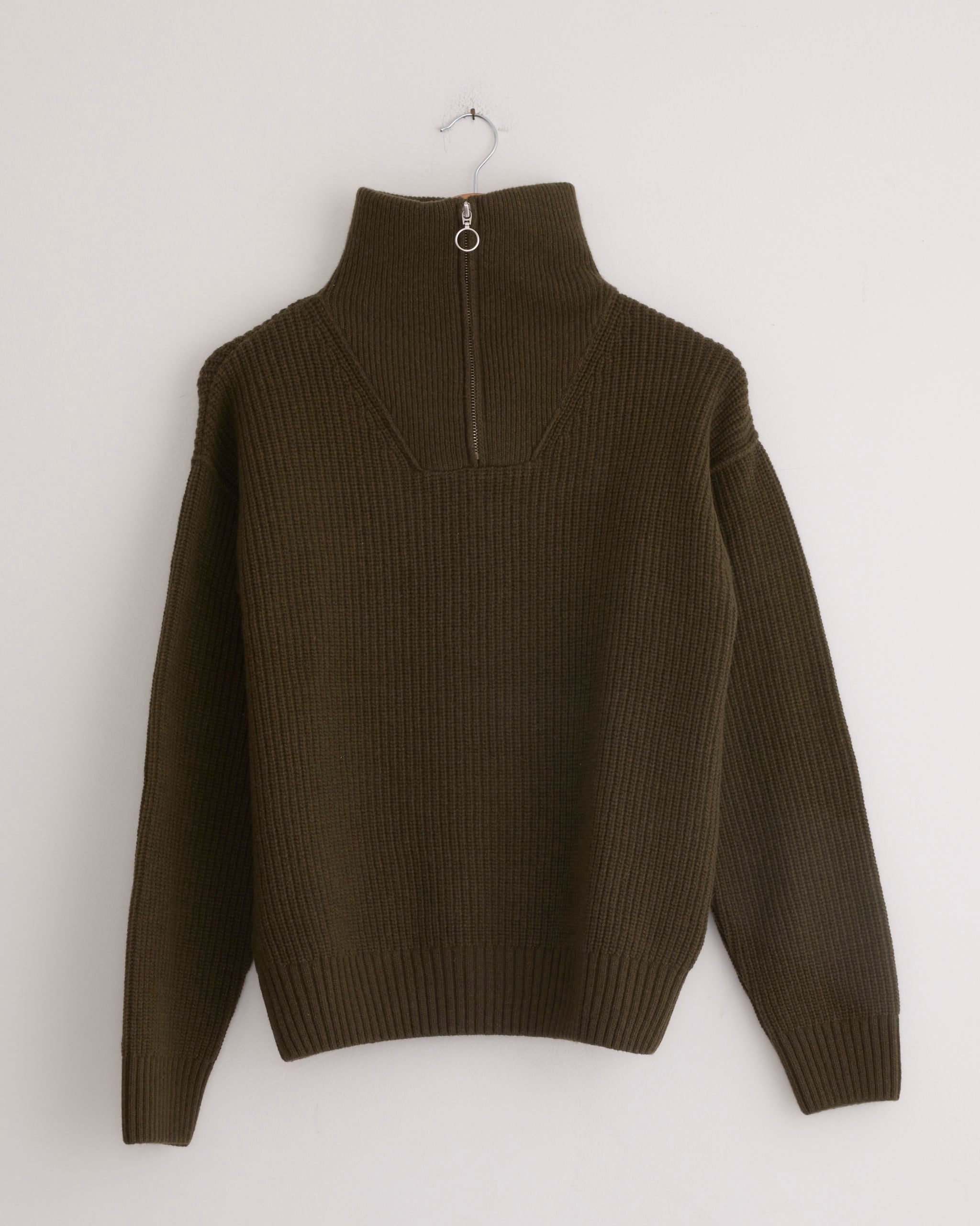 The Fly Deck Sweater in Canteen by Girls of Dust, made from ribbed merino wool with long sleeves and a high zip collar, hangs on a simple metal hook against a plain light background.